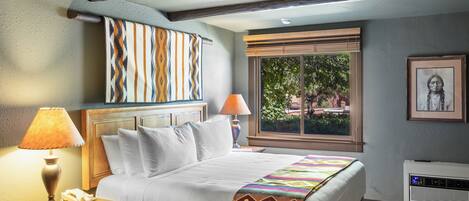 A hotel room with a bed, bedside lamps, a window with blinds, a wall-mounted air conditioner, and a framed picture.