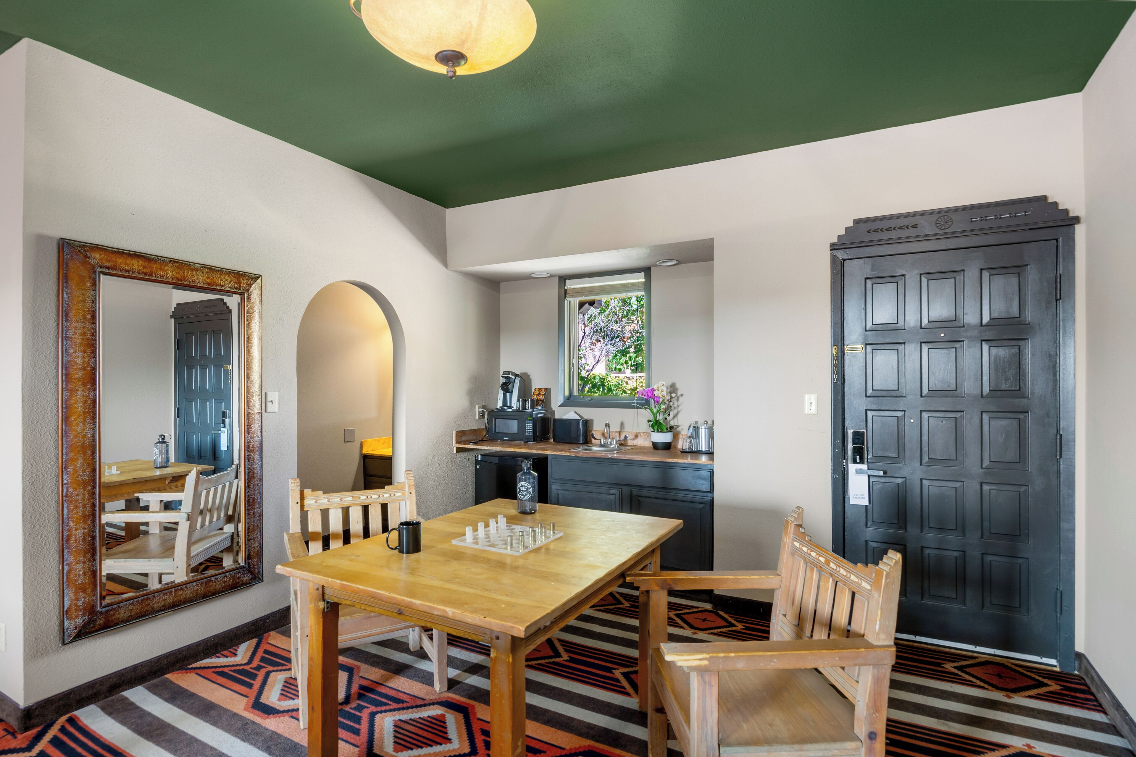 Un comedor con una mesa y sillas de madera, un tablero de ajedrez, un espejo grande, una zona de cocina con ventana y una puerta oscura.