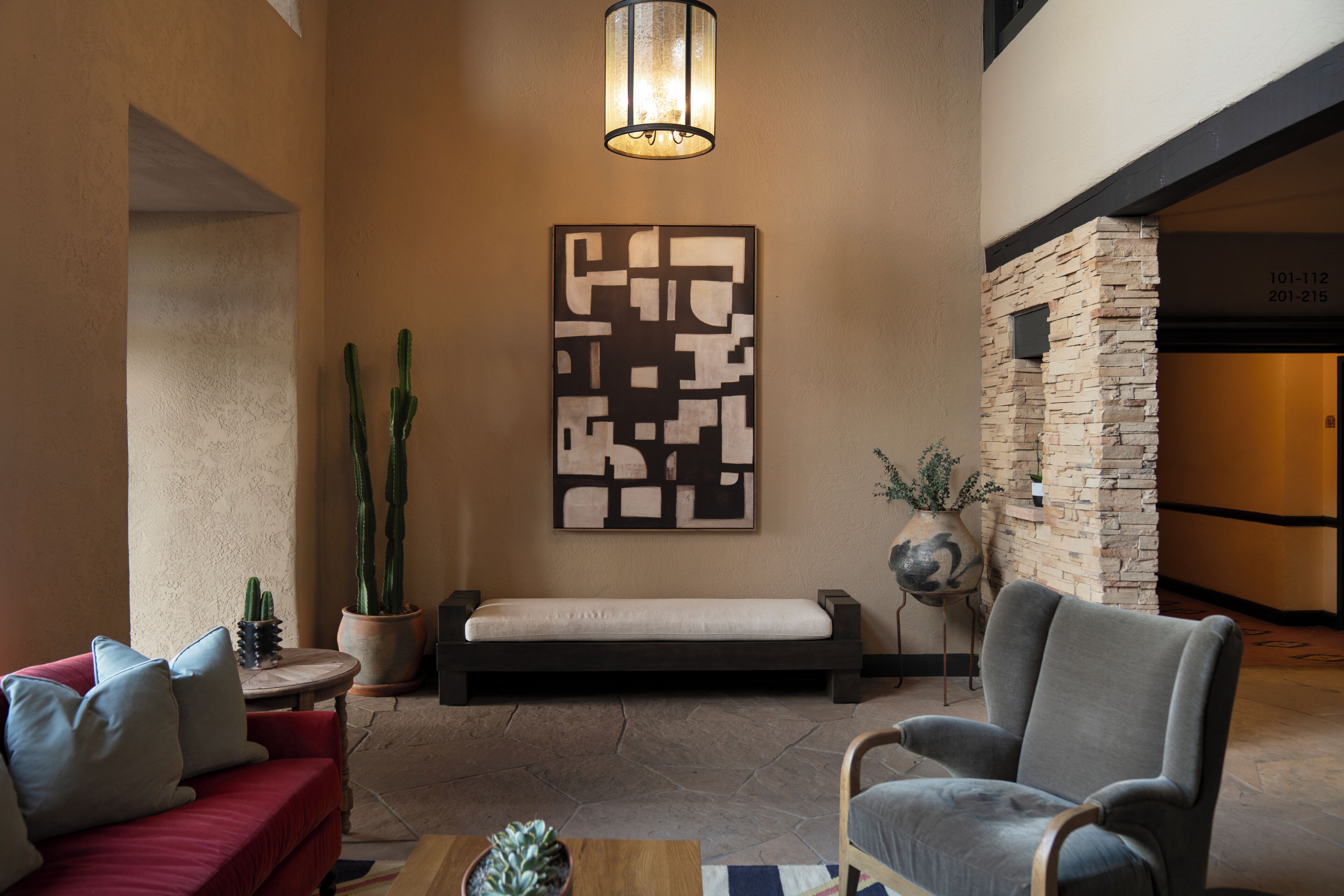 A lobby with a red sofa, a grey armchair, a wall-mounted light fixture, a cactus, and a geometric wall art piece.
