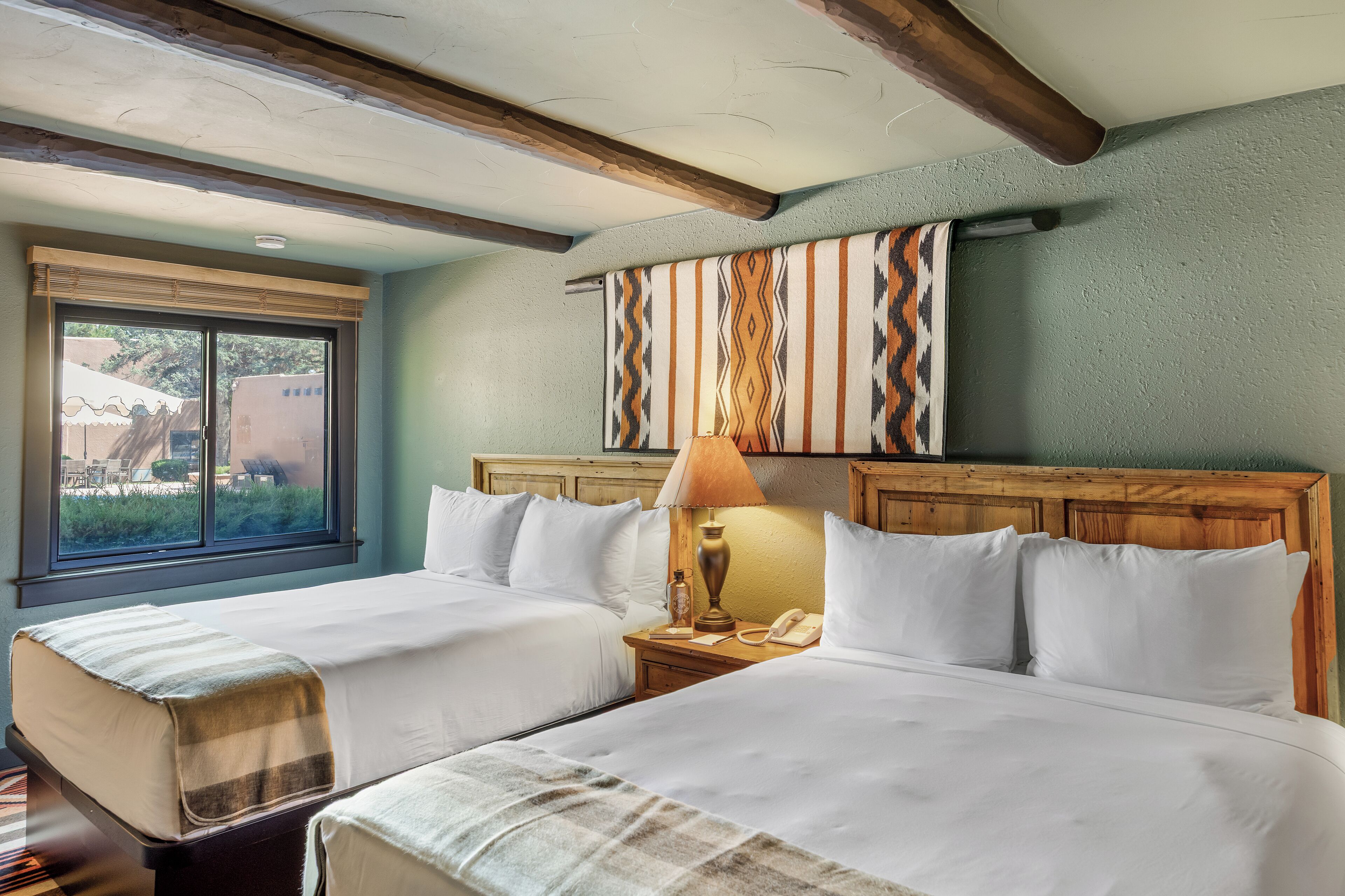 A hotel room with two beds, a window with a view, a wooden headboard, and a wall-mounted lamp.