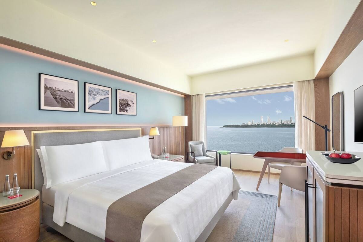 Une chambre d’hôtel avec un grand lit, un bureau, une chaise et une vue sur les toits d’une ville, au-delà d’une étendue d’eau.