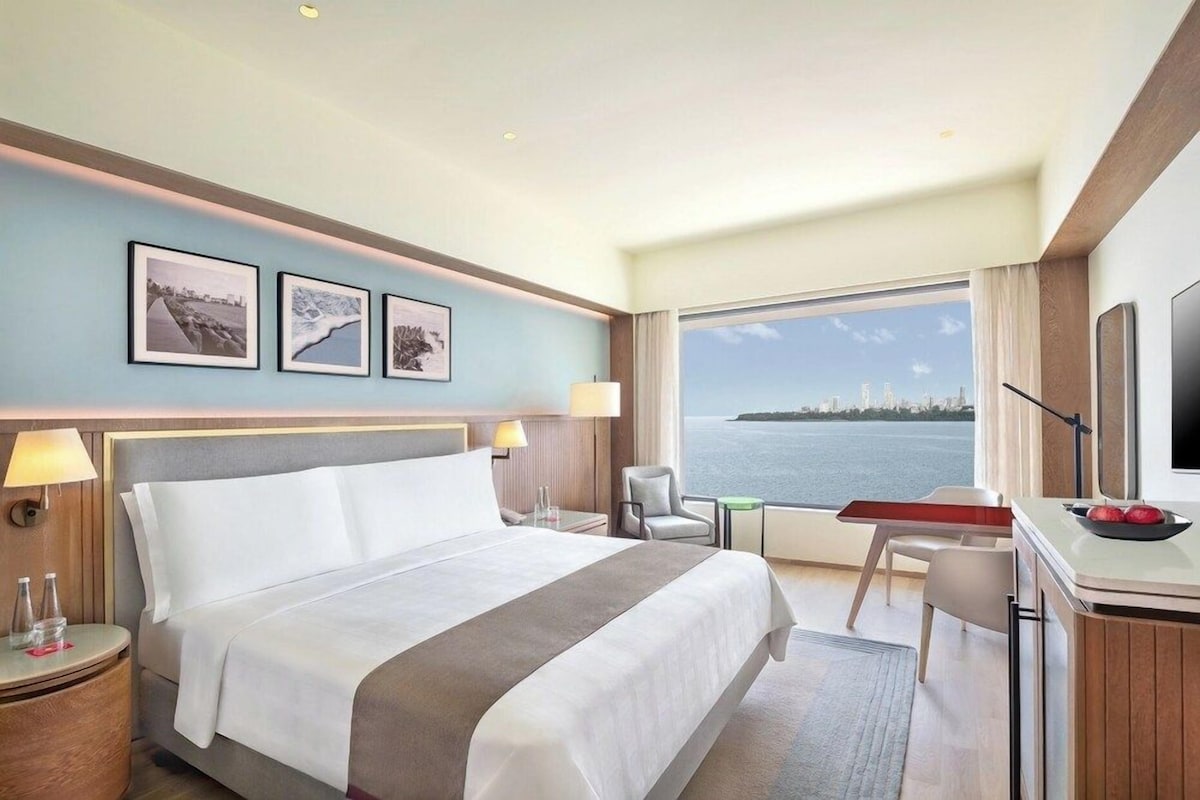 Habitación de hotel con cama, escritorio, silla y un amplio ventanal con vista al mar.