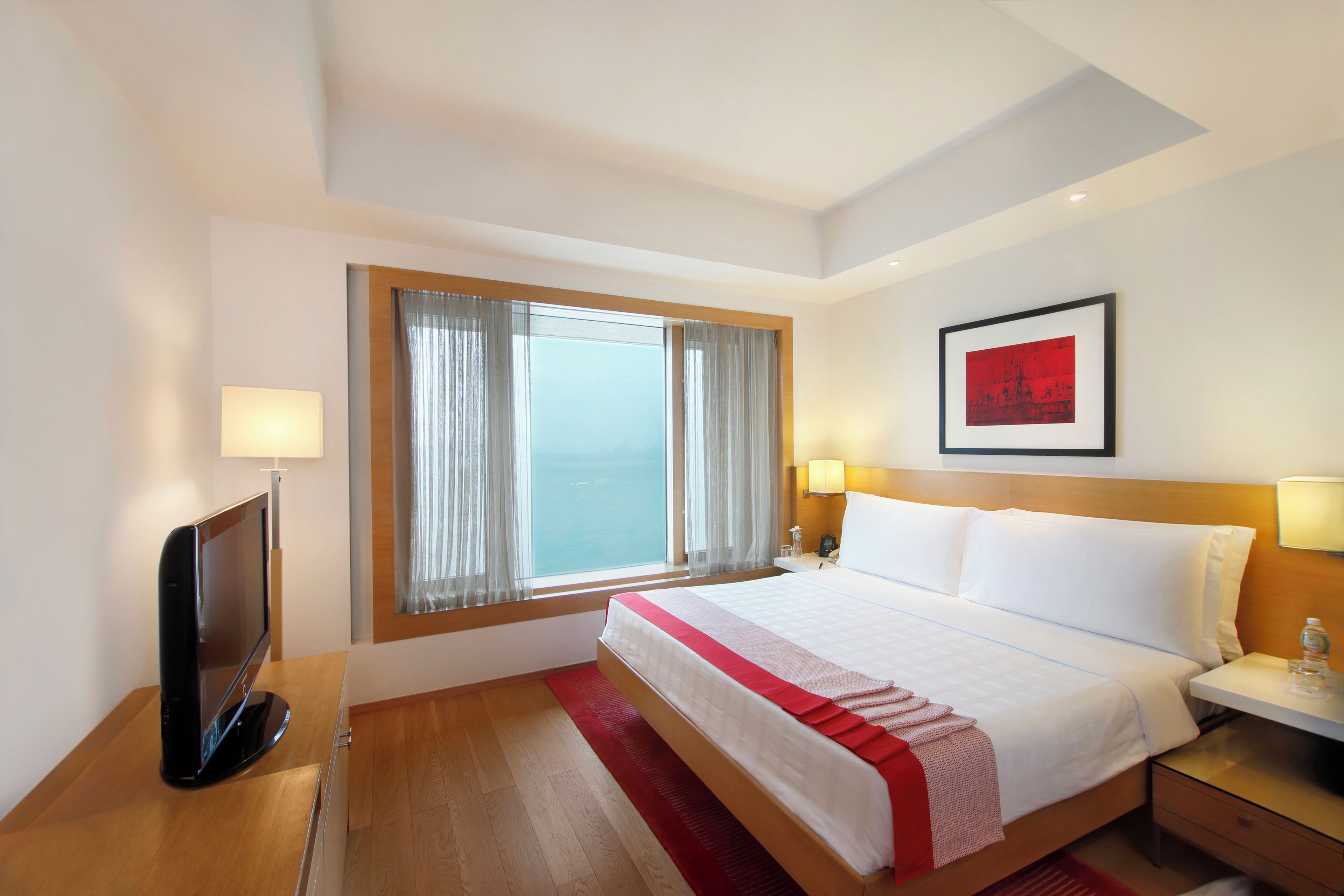 Una habitación de hotel con cama, televisión, ventana con cortinas, mesita de noche y un cuadro en la pared.
