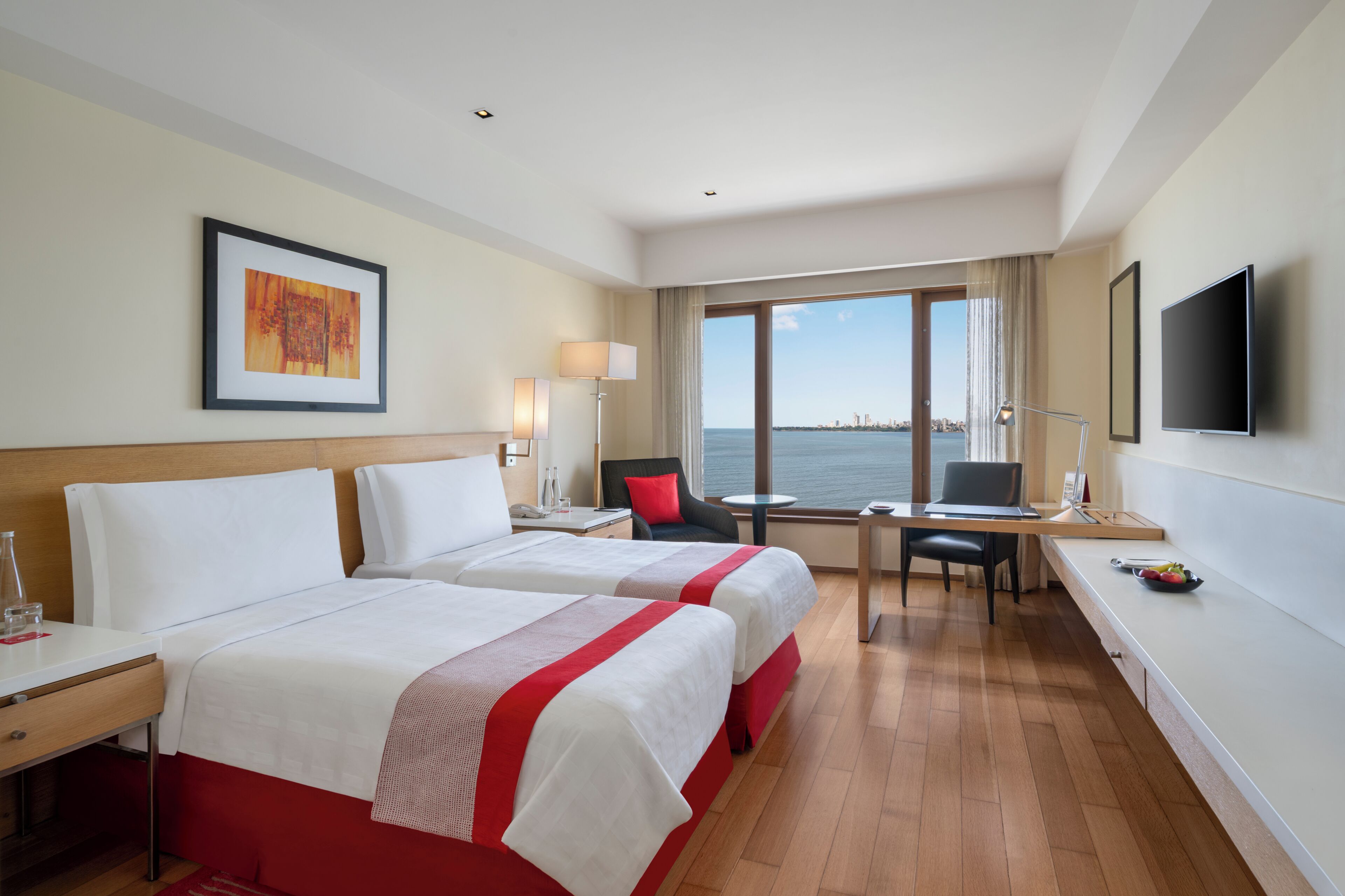 A hotel room with two beds, a desk, a chair, a TV, and a large window with a view of the sea.