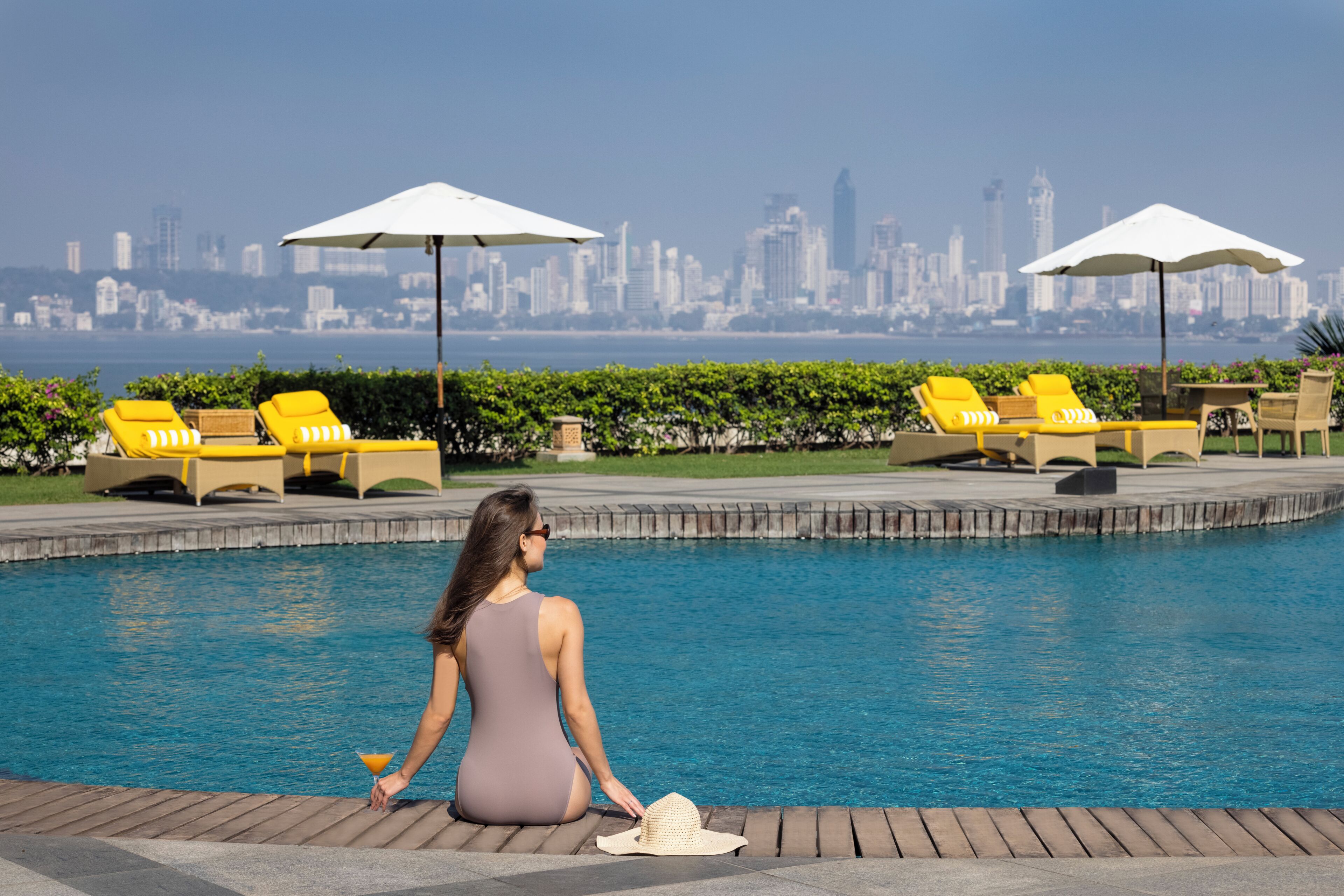 A poolside area with lounge chairs and umbrellas, a city skyline in the background, and a person sitting by the pool.