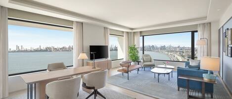 A modern hotel room with a city view, a flat-screen TV, a dining area, and a seating area with a sofa and armchairs.