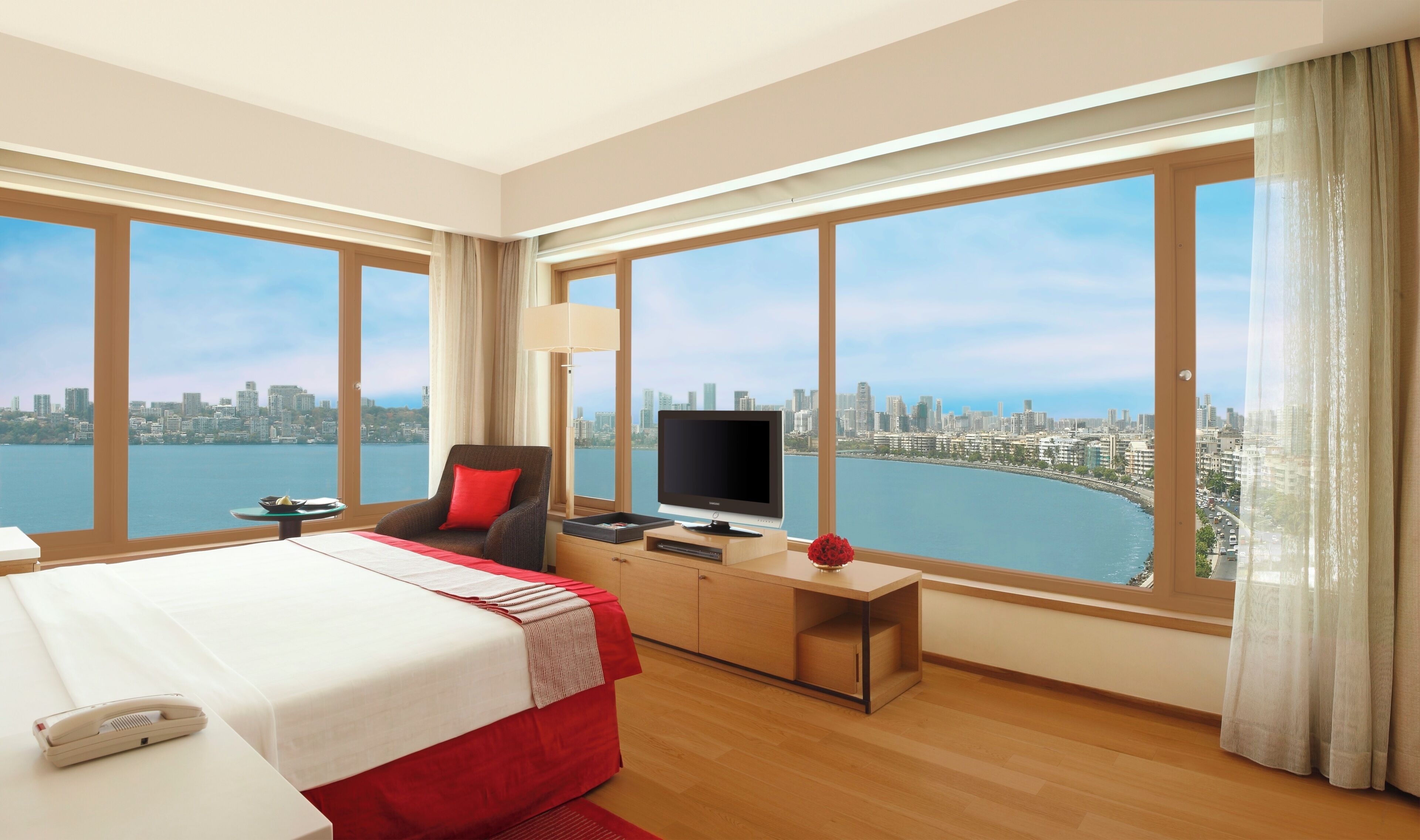 A hotel room with a large bed, a TV, a desk, and a view of a city skyline and water.