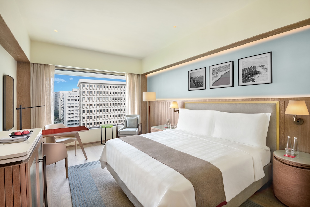 A hotel room with a bed, desk, chair, and a view of buildings.