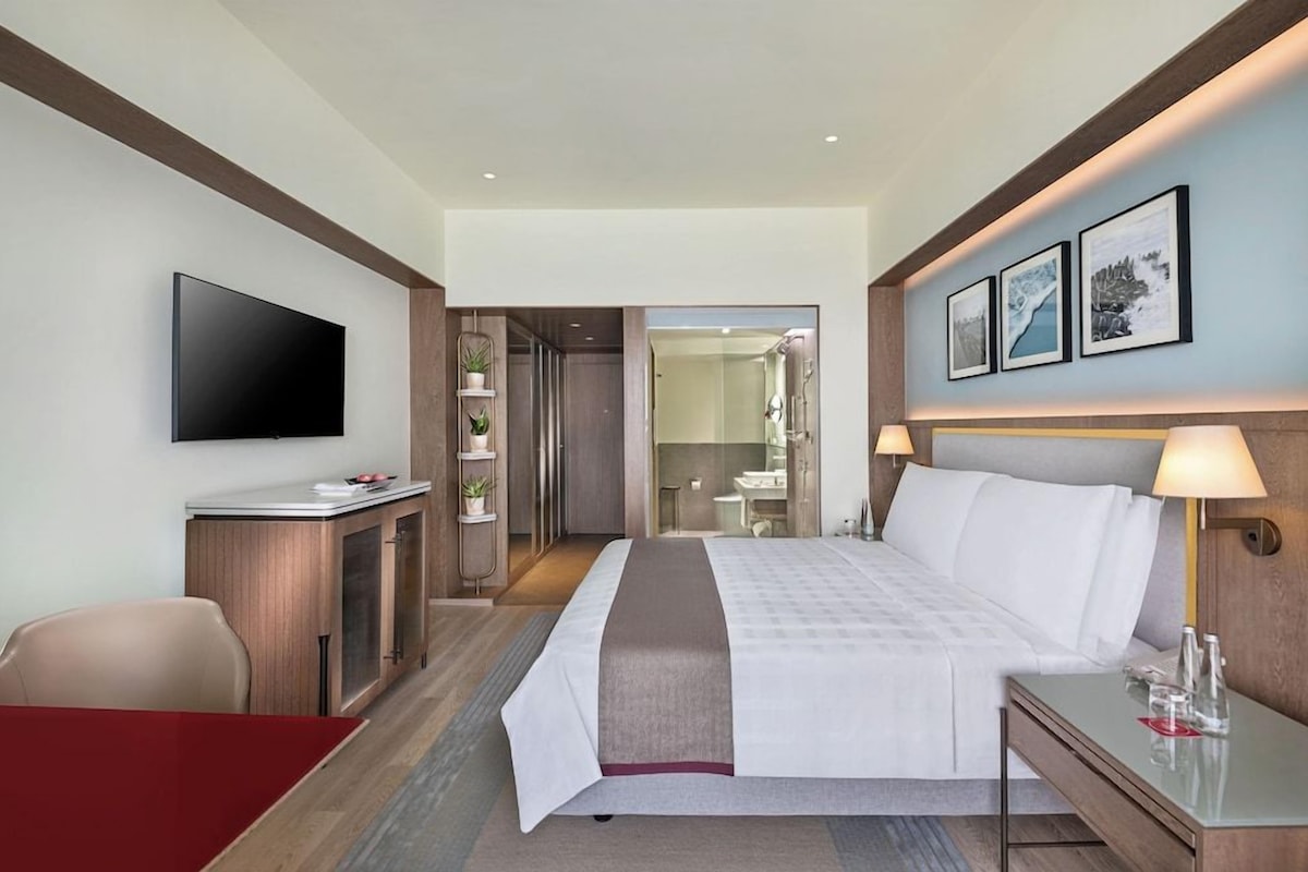 A hotel room with a bed, bedside table, TV, desk, chair, and artwork on the wall.
