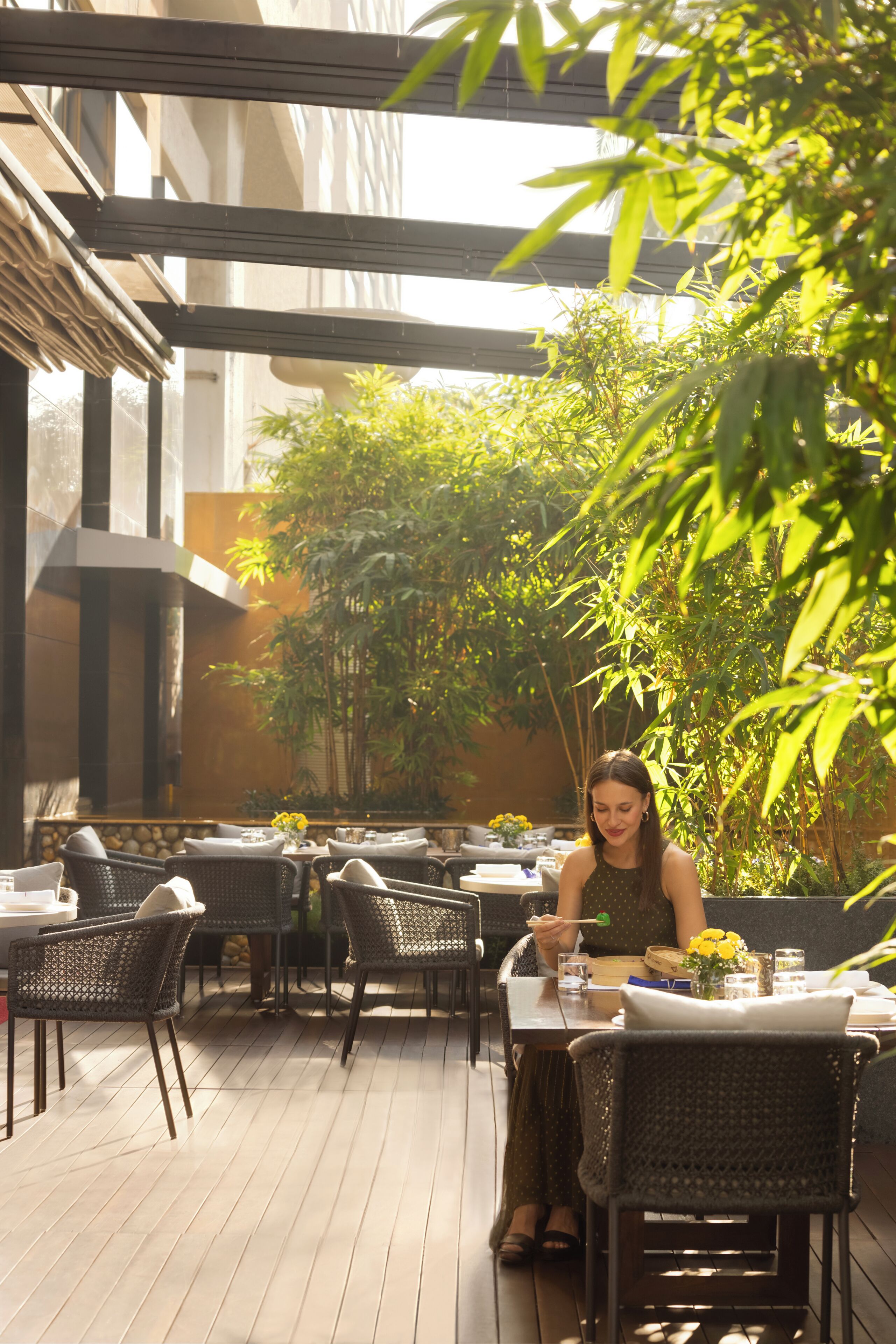An outdoor dining area with tables and chairs, surrounded by greenery and a building structure.