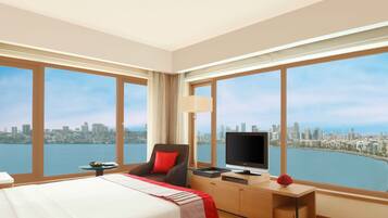 A hotel room with a large bed, a desk with a TV, a chair, and a view of a city skyline and water.