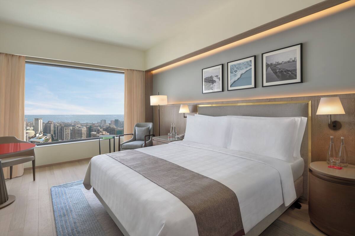 Une chambre d’hôtel avec un grand lit, un bureau, une chaise, une vue sur la ville et deux tableaux encadrés au mur.