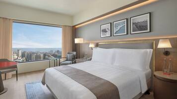 A hotel room with a large bed, a desk, a chair, a view of the city, and two framed pictures on the wall.