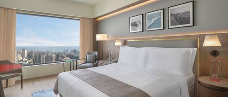 A hotel room with a large bed, a desk, a chair, a view of the city, and two framed pictures on the wall.