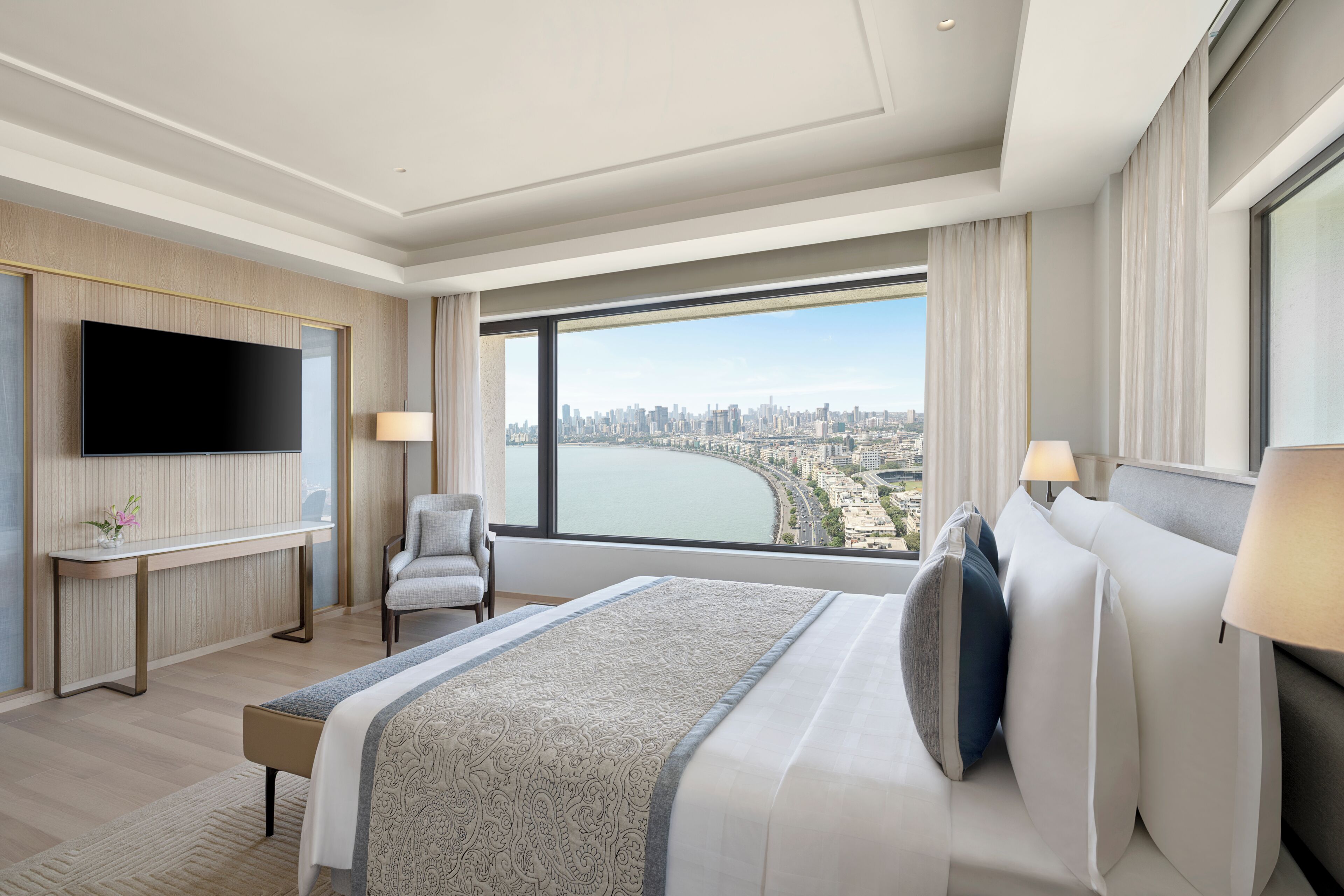 A modern hotel room with a large bed, a flat-screen TV, a desk, and a large window offering a city view.