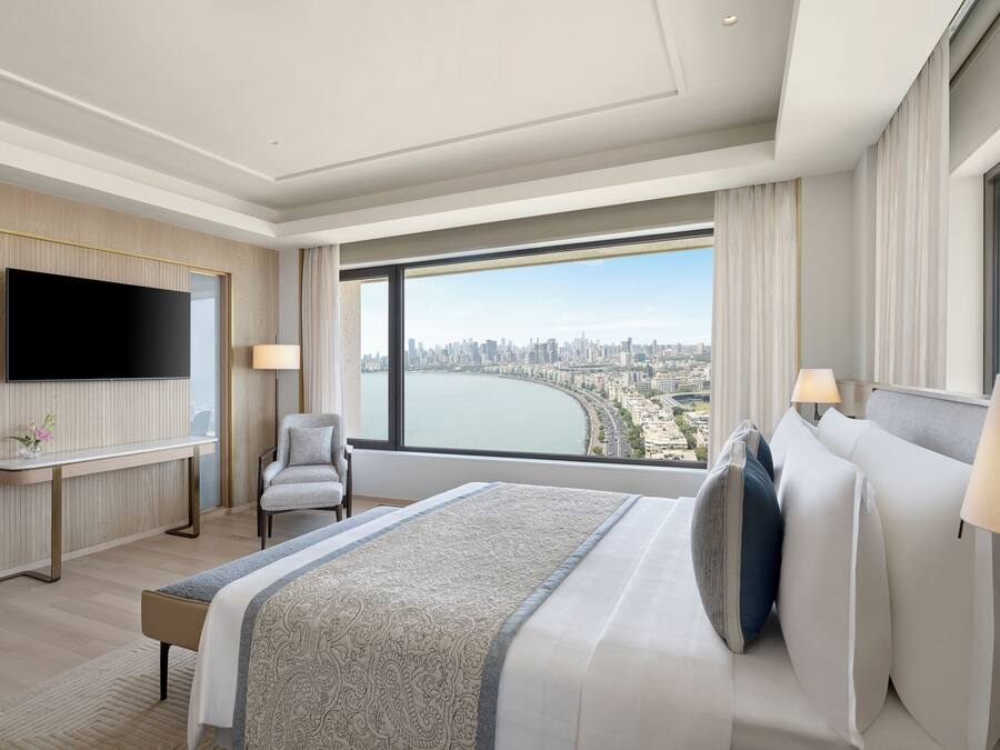 A modern hotel room with a large bed, a flat-screen TV, a desk, and a large window offering a city view.