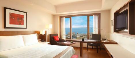 A hotel room with a large bed, a desk with a chair, a TV, and a window with a city view.