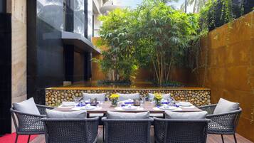Un espace extérieur aménagé pour les repas, avec un mur en pierre, des plantes en bambou et une vue sur un bâtiment.