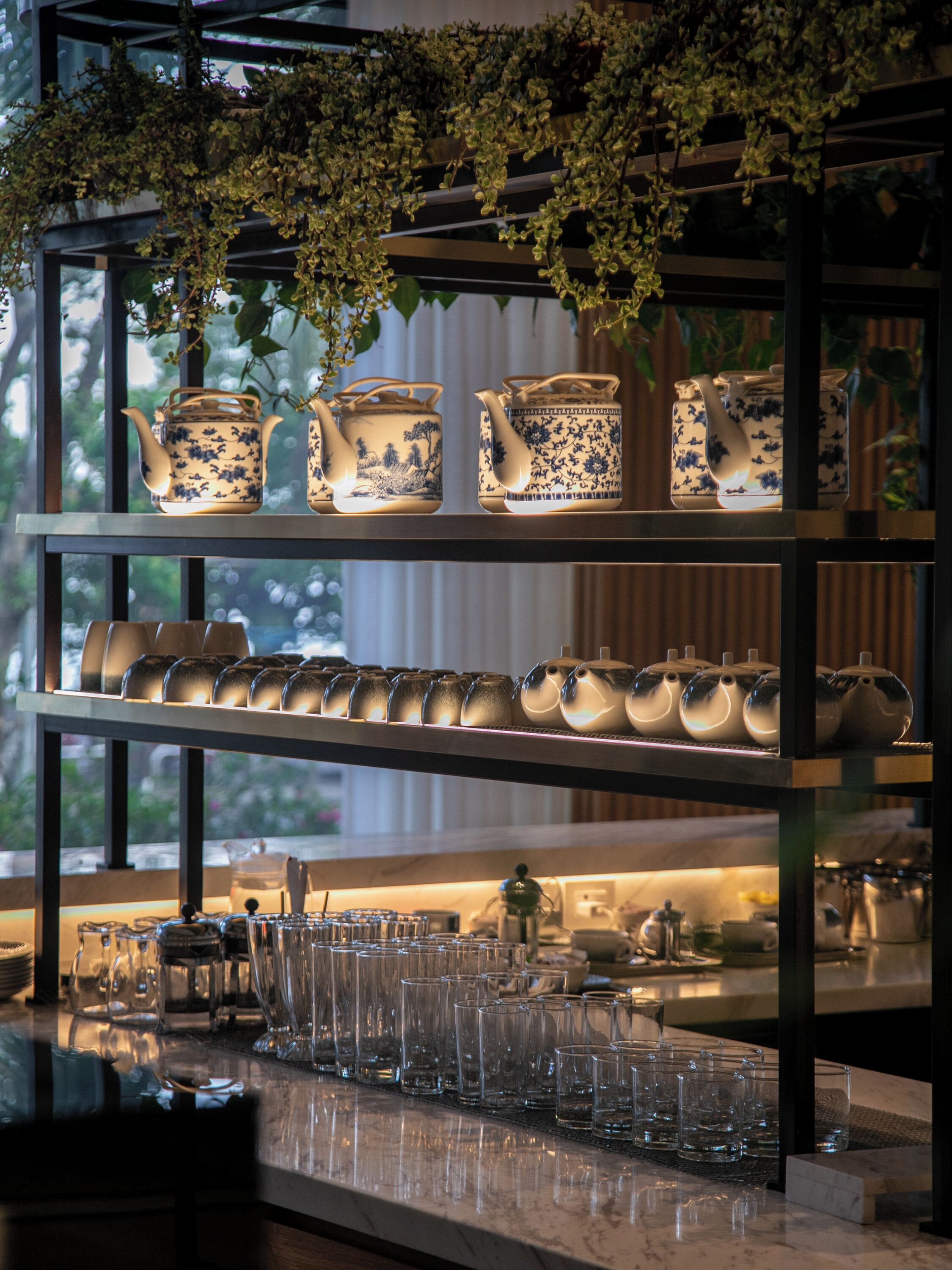 Una cocina con estantes que exhiben teteras y tazas, una encimera con vasos y una ventana con follaje.