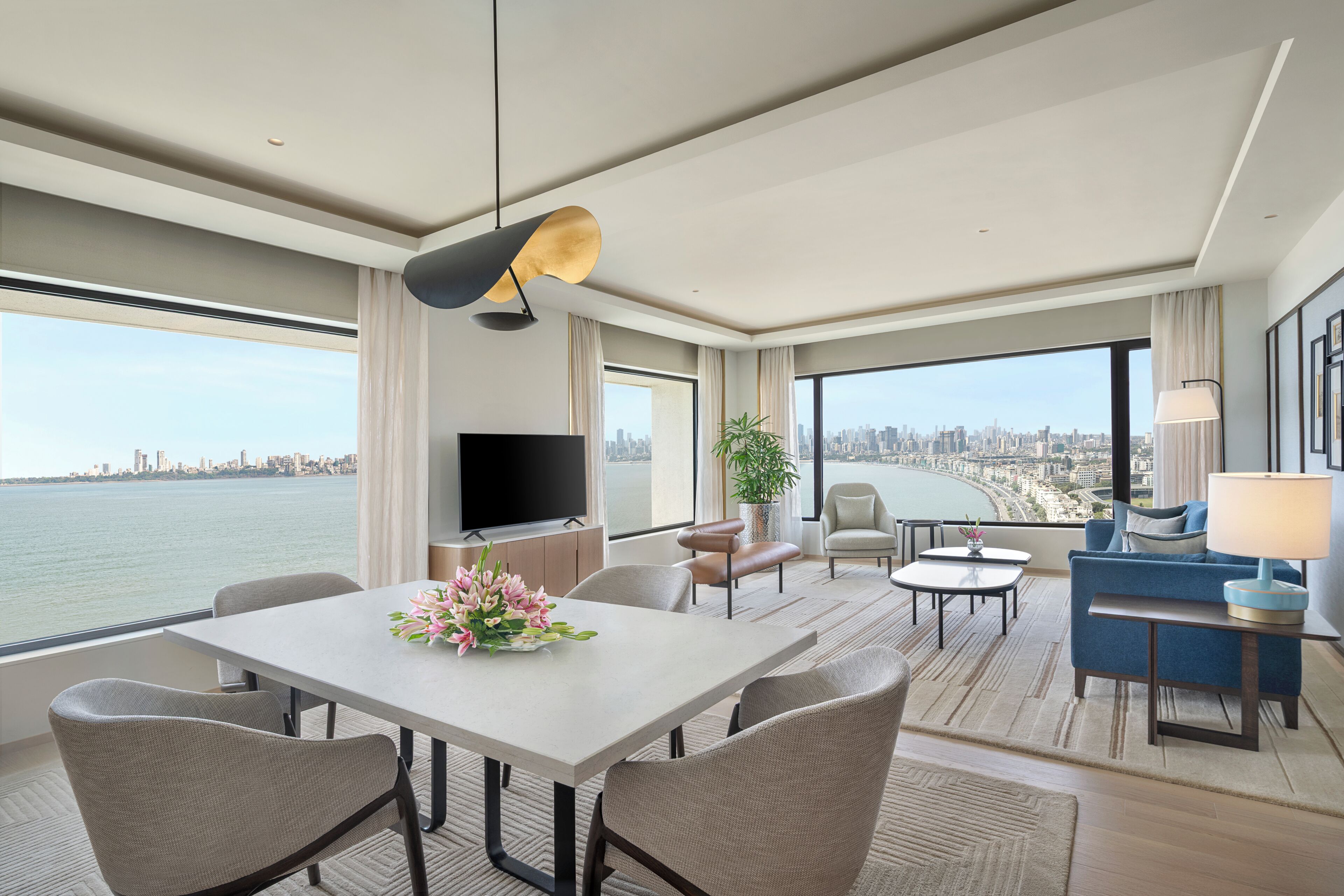 A modern hotel room with a dining area, seating, a TV, and a large window with a city view.