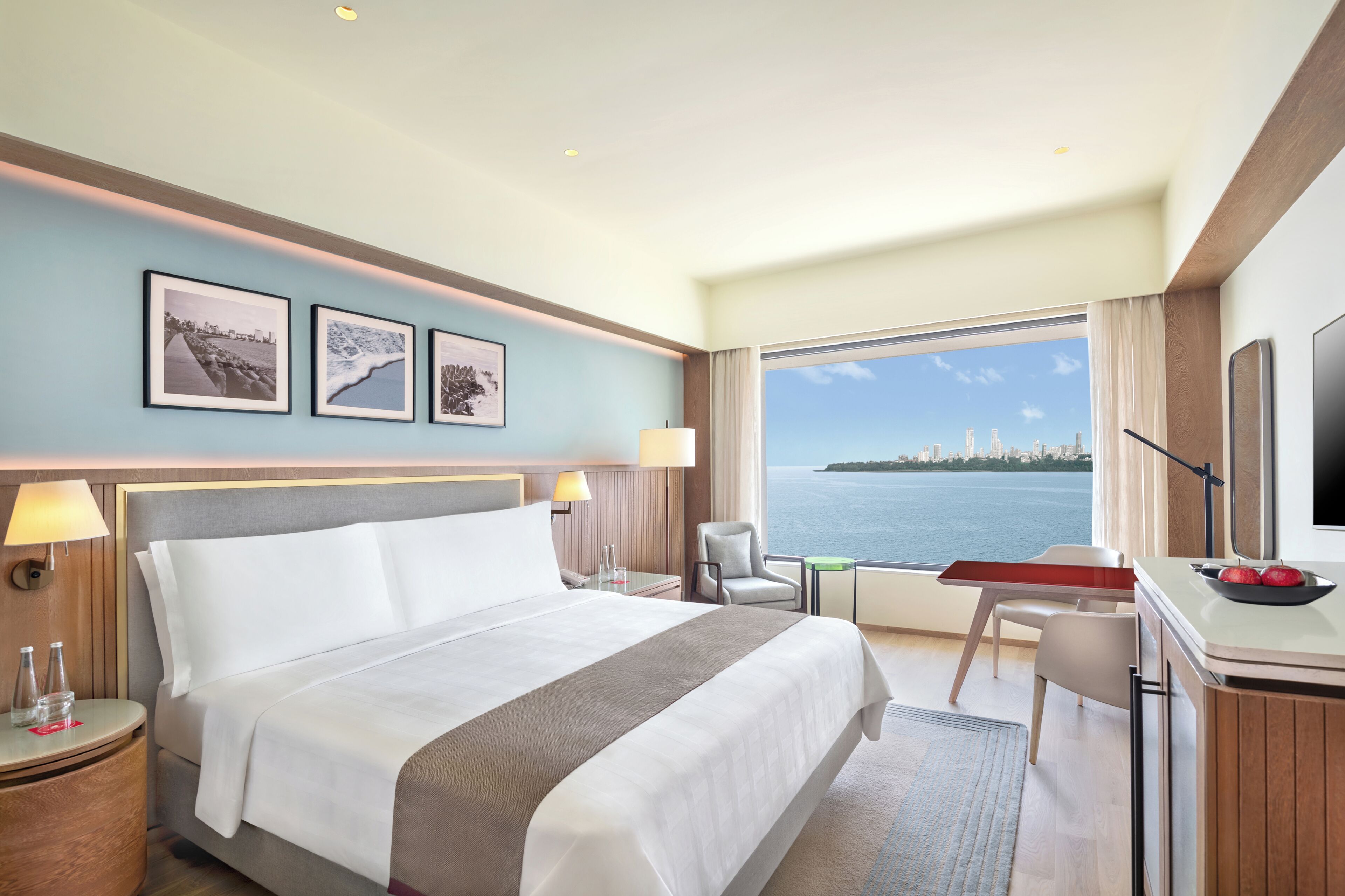 A hotel room with a bed, desk, chair, and a large window with a city view.