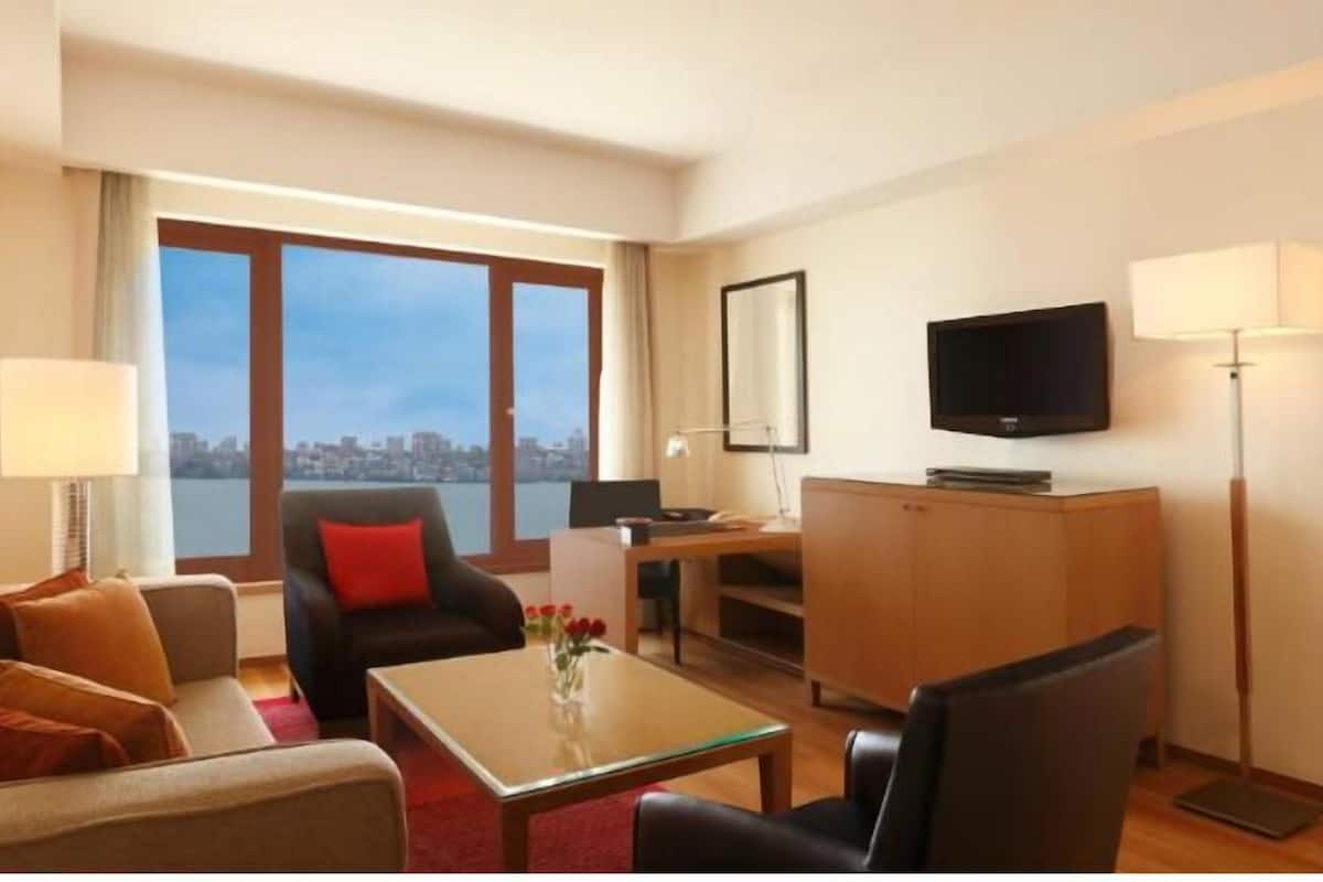 Une chambre d’hôtel équipée d’un canapé, d’un fauteuil, d’une table basse, d’un bureau, d’une télévision et d’une grande fenêtre donnant sur la ville.