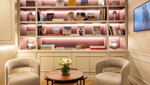 Uma estante bem organizada com vários livros e objetos decorativos, uma mesa de madeira redonda com um vaso de flores e duas poltronas estofadas.