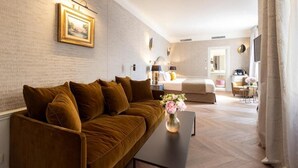 Quarto de hotel moderno com um sofá marrom, uma cama, uma mesinha com um vaso de flores, e um banheiro visível ao fundo.