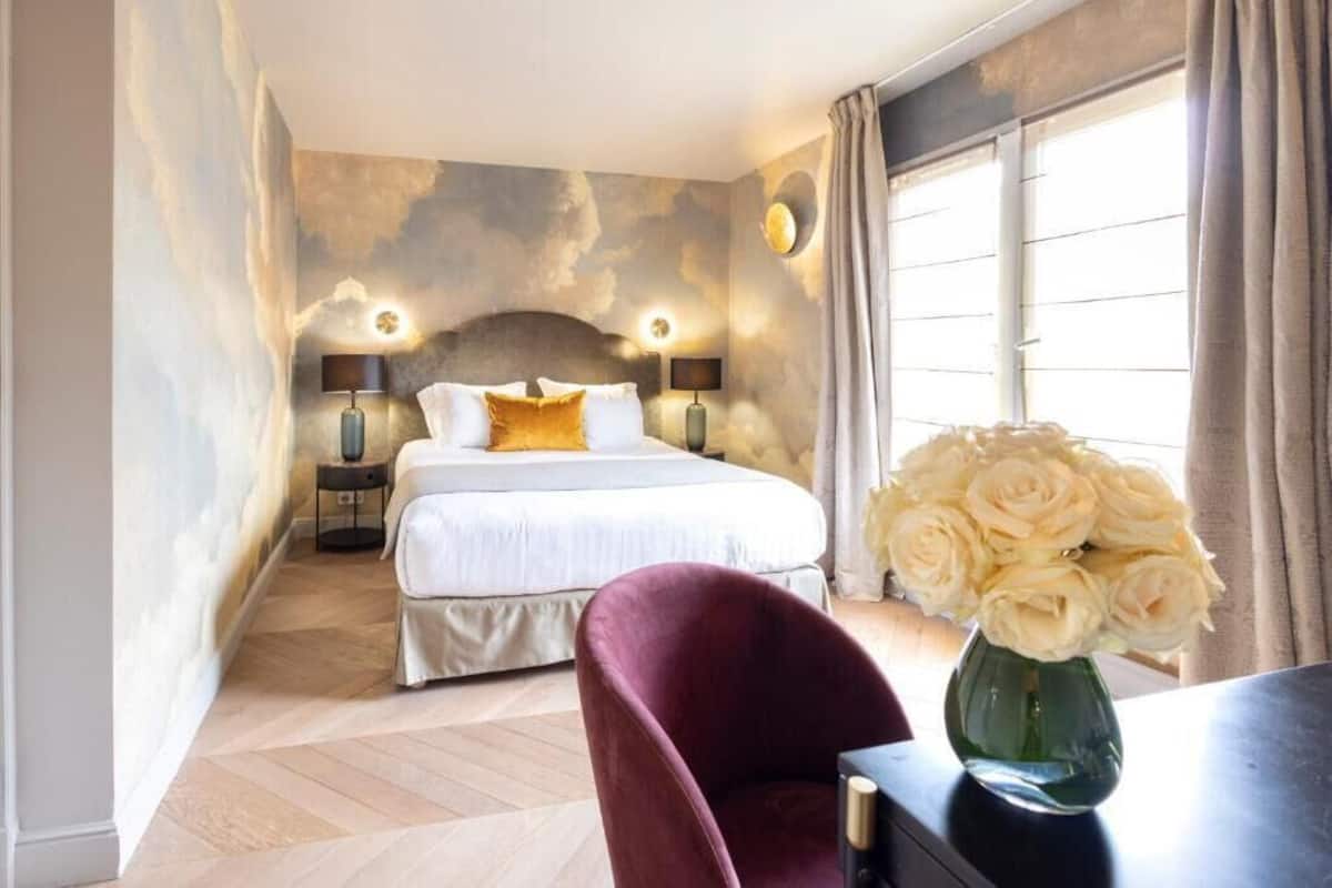 Une chambre à coucher avec un lit, des lampes de chevet, un bureau agrémenté d’un vase de fleurs et une fenêtre avec des rideaux.