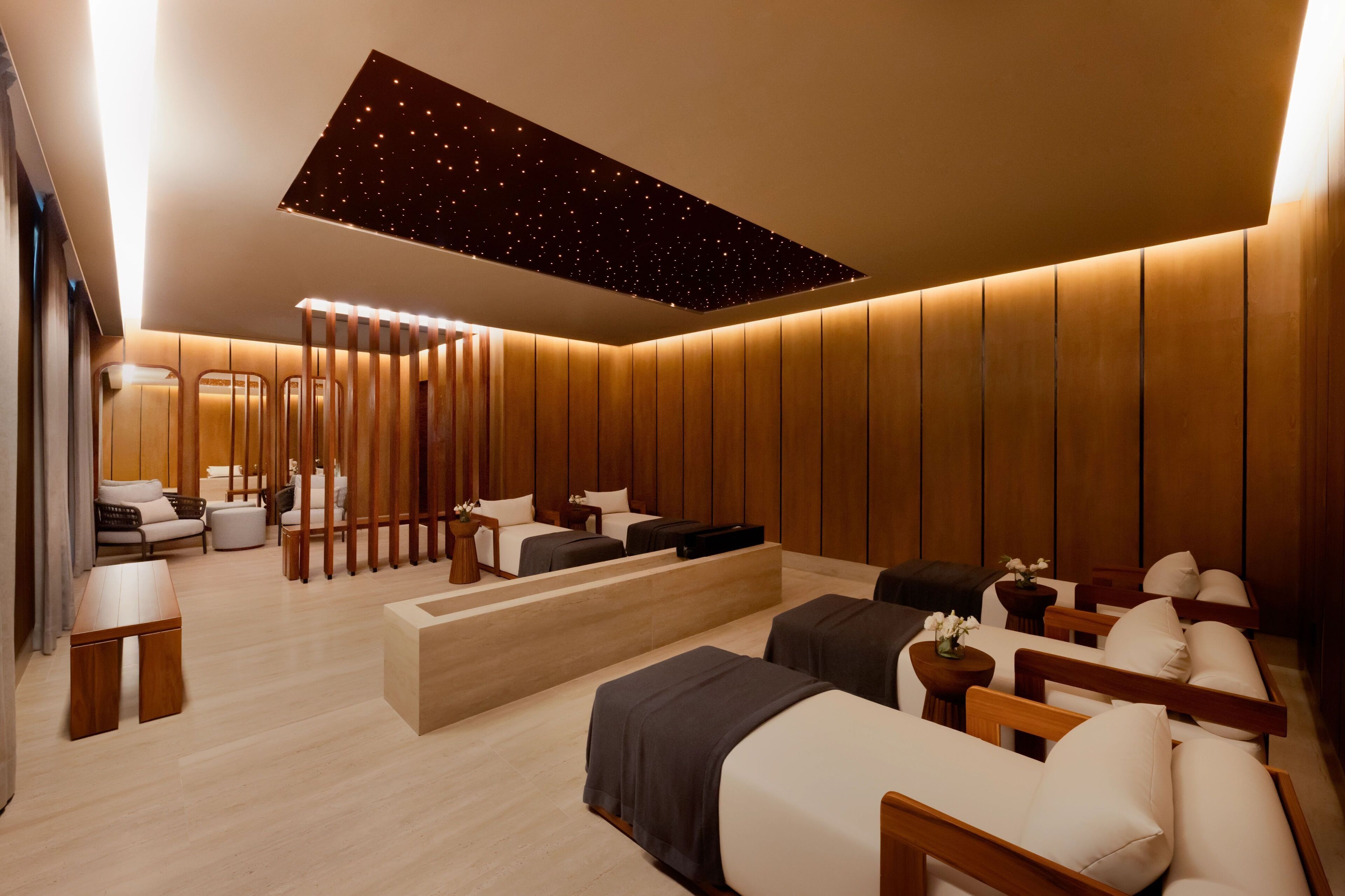 A modern spa room with a starry ceiling, wooden panel walls, and a variety of seating options including a sofa, chairs, and a massage table.