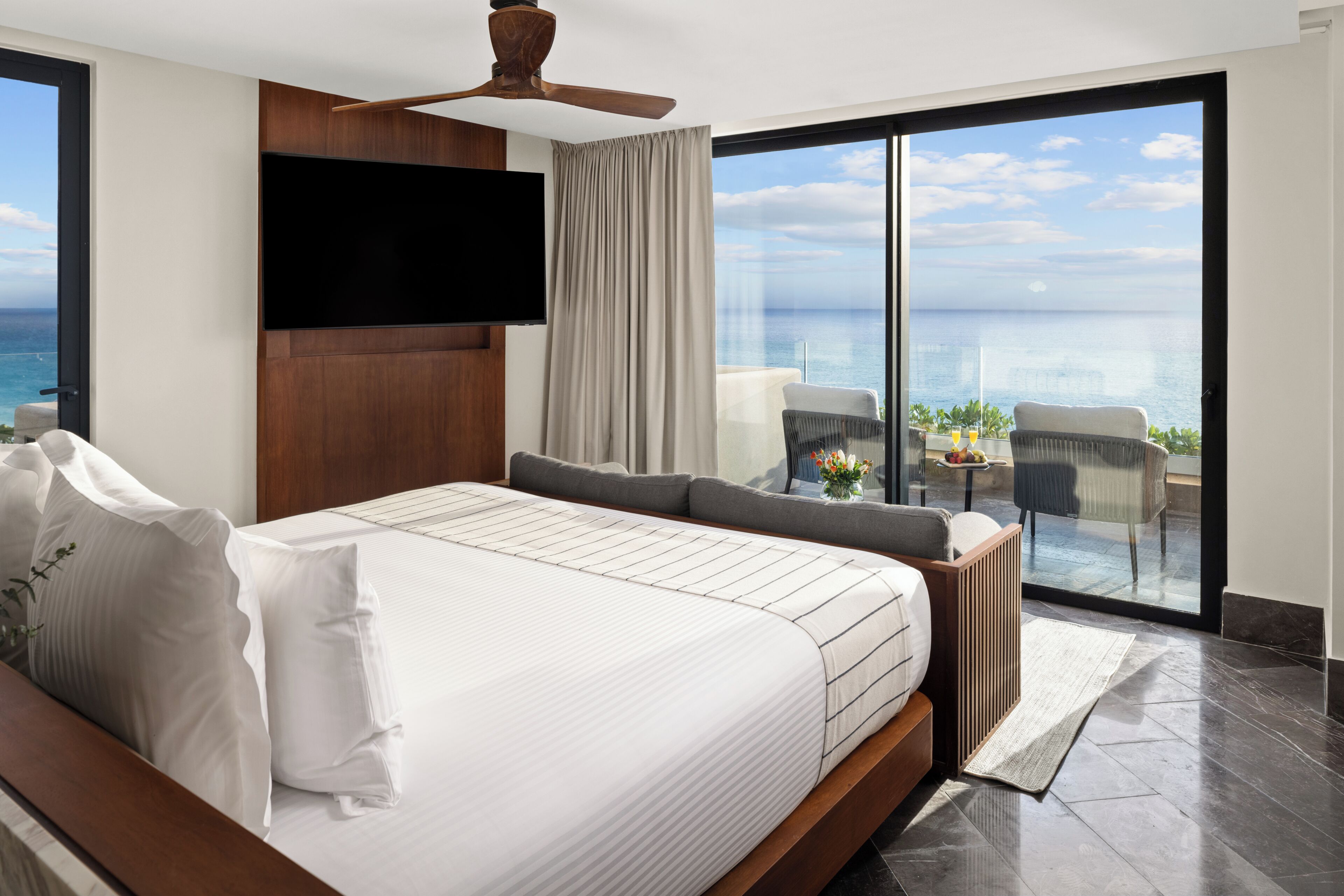 Ein modernes Hotelzimmer mit einem großen Bett, einem Flachbildfernseher, einem Deckenventilator, einem Balkon mit Meerblick und einer Sitzecke mit Tisch und Stühlen.