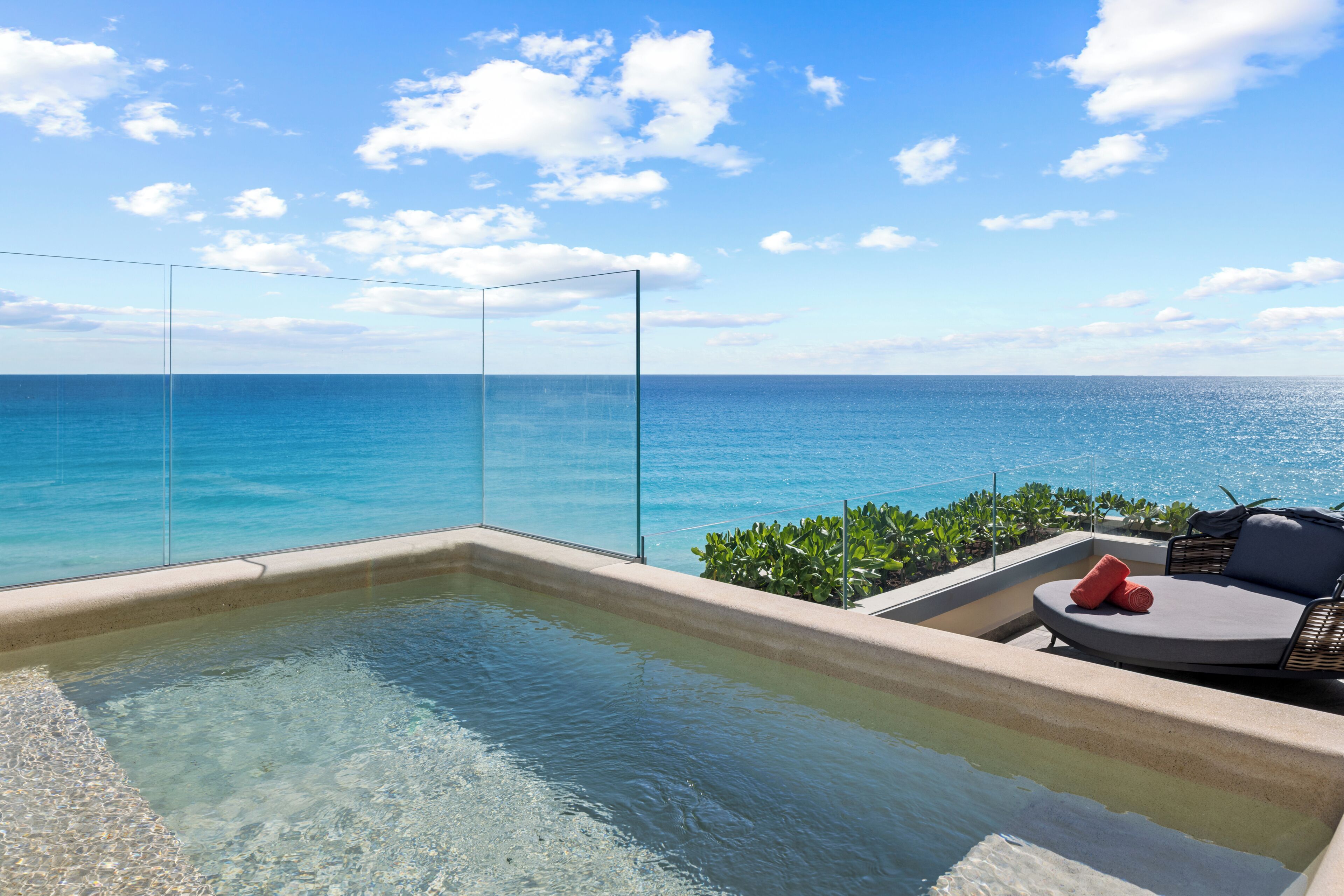 A clear view of the ocean from a glass-enclosed infinity pool with a lounge area and a hot tub.