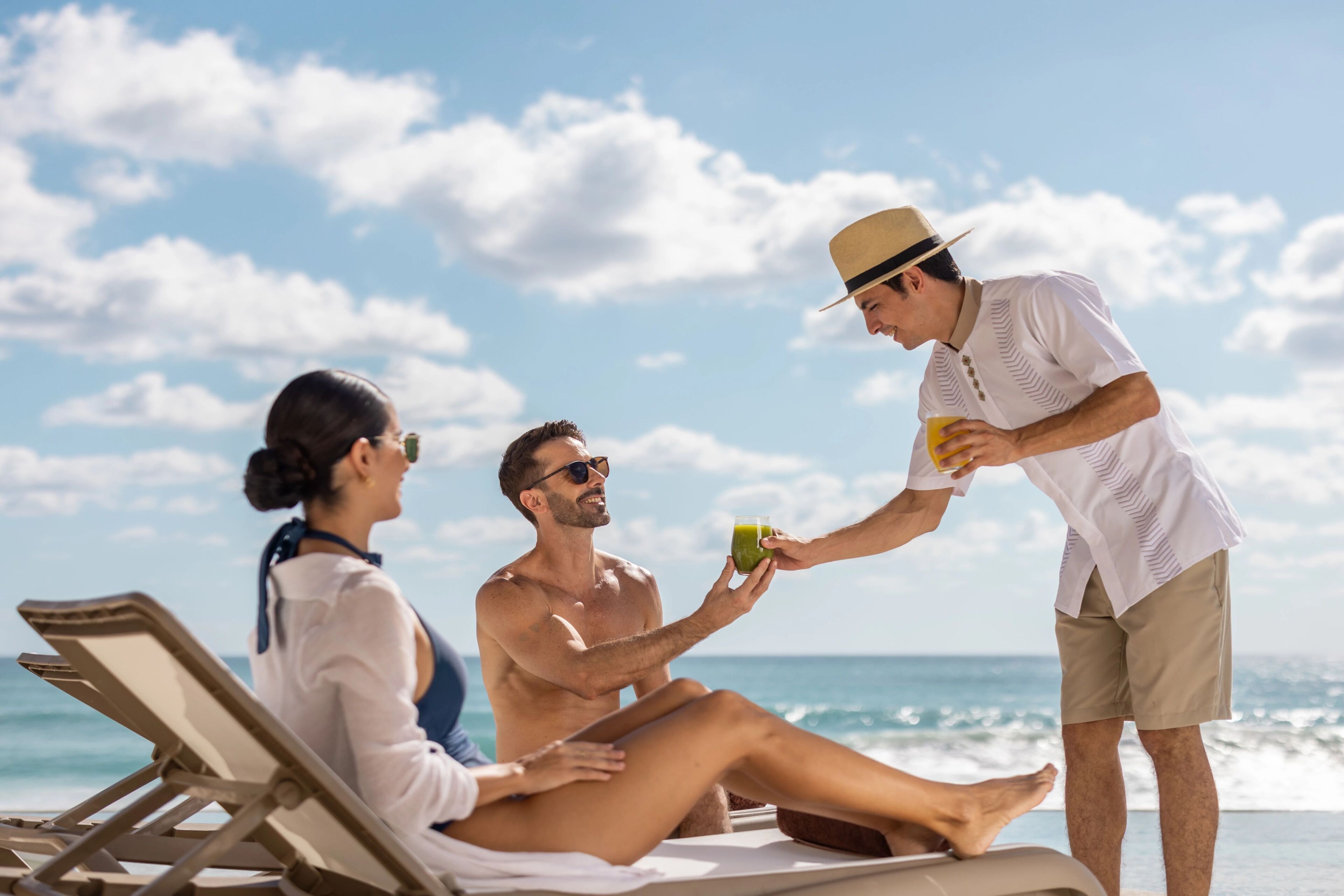 A beach scene with a man and a woman sitting on a lounge chair, and another person standing nearby.