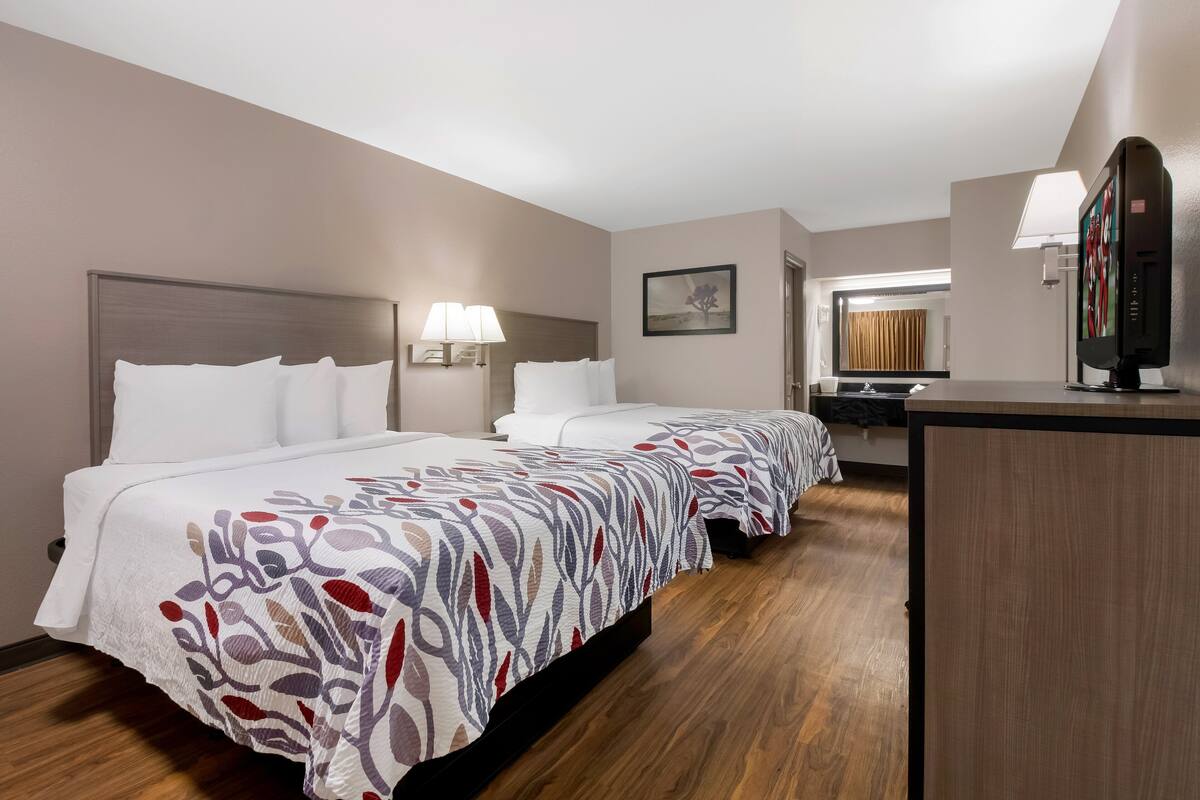 A hotel room with two beds, a desk, a TV, and a bathroom visible through an open door.