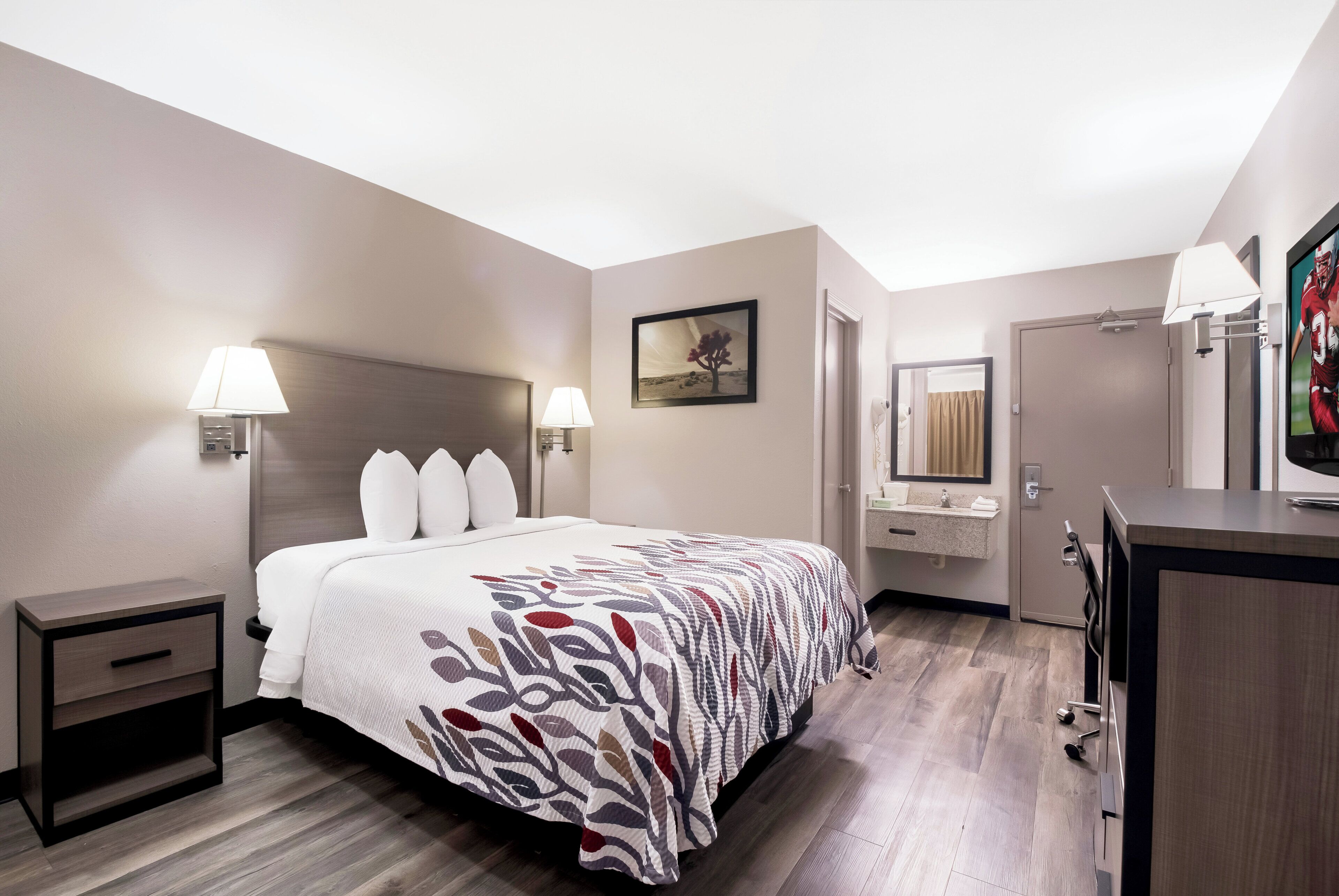 A hotel room with a bed, bedside lamps, a painting, a bathroom with a sink and mirror, and a TV.