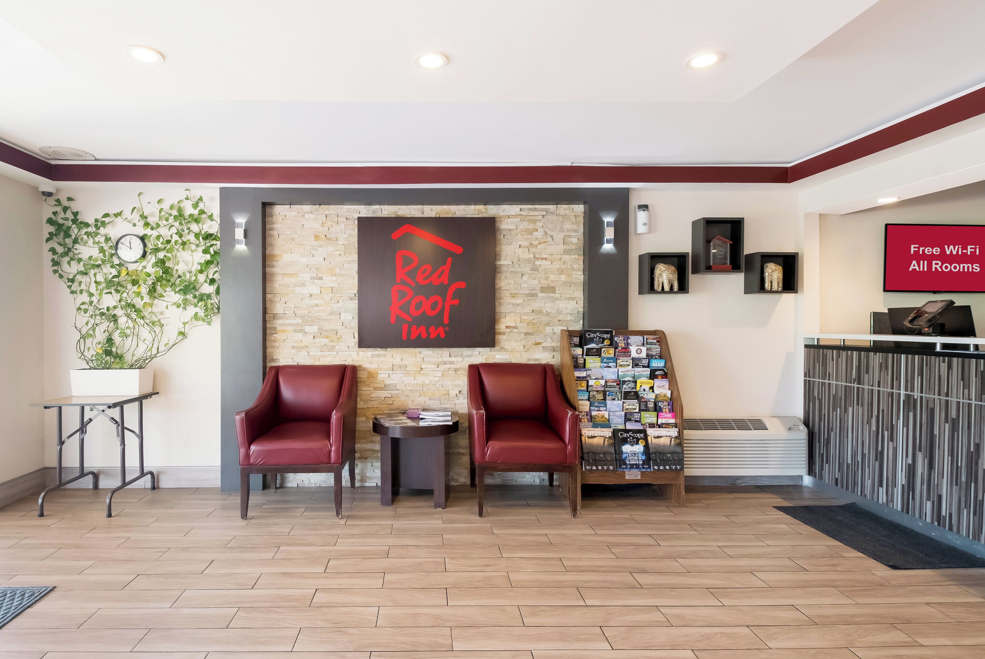A reception area with a Red Roof Inn sign, two leather chairs, a small table, a wall-mounted clock, a bulletin board with various items, and a sign indicating free Wi-Fi in all rooms.
