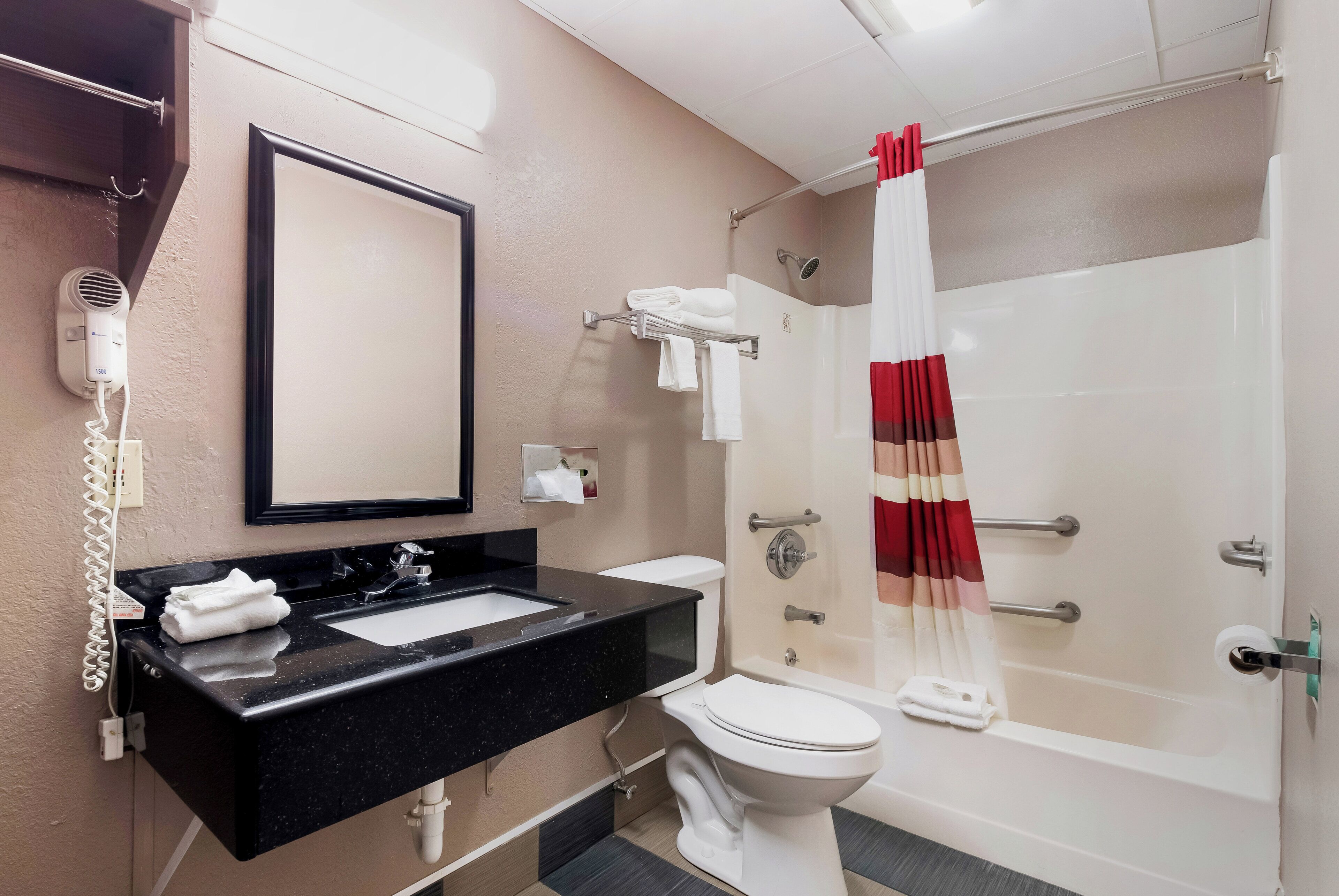 A bathroom with a black countertop, a white sink, a toilet, a shower with a red and beige curtain, and a hairdryer mounted on the wall.