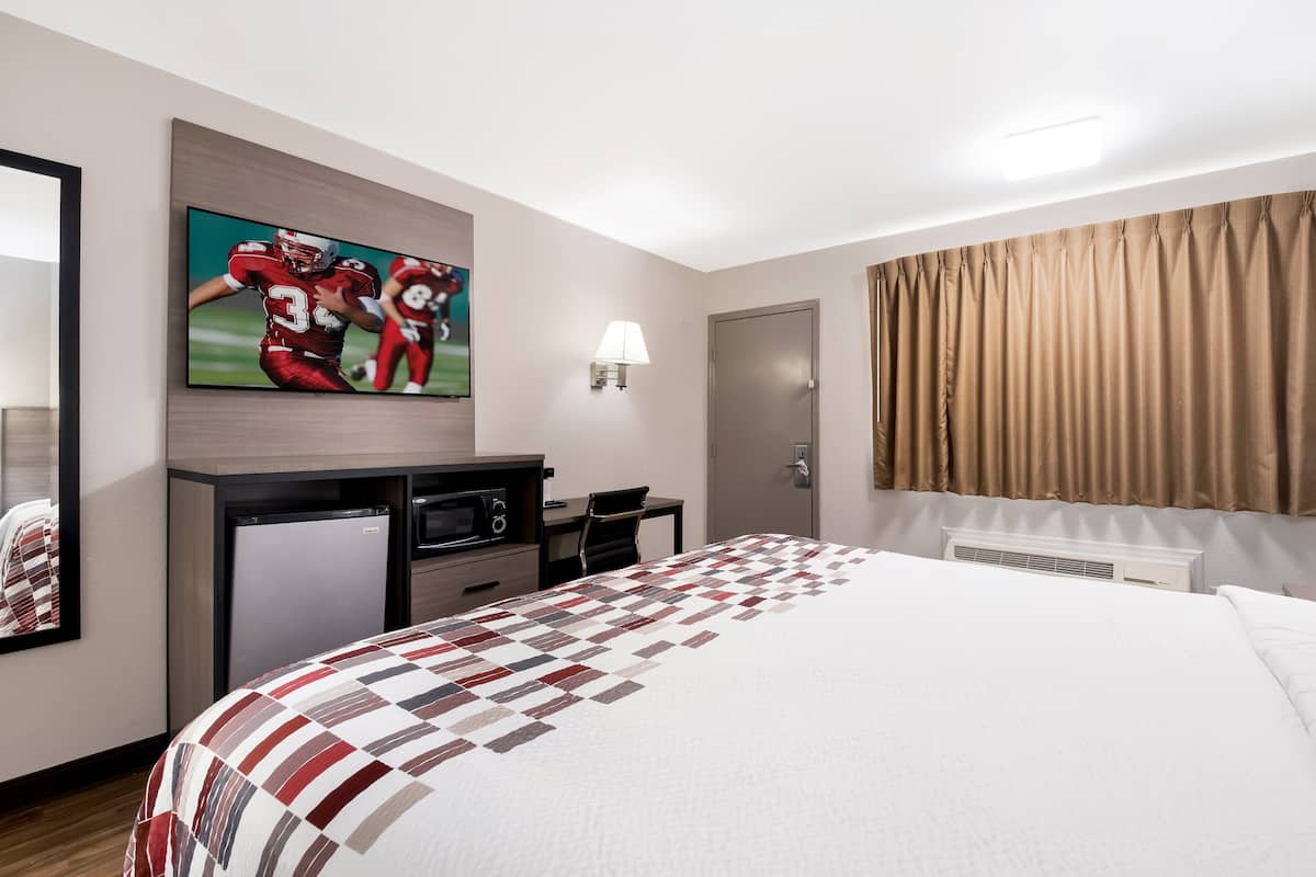 A hotel room with a large bed, a flat-screen TV mounted on the wall, a desk with a chair, a small refrigerator, and a window with curtains.