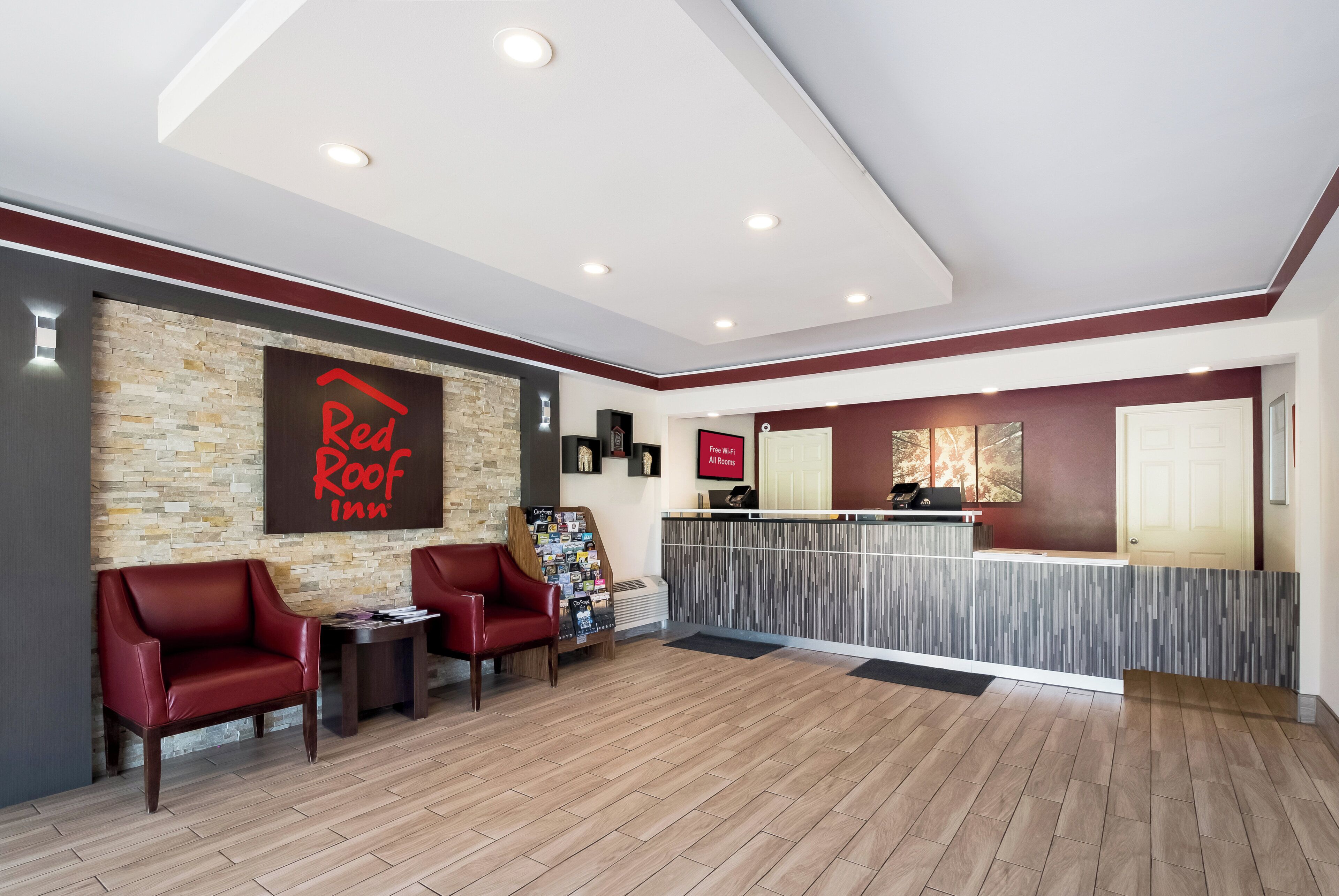 A Red Roof Inn reception area with a stone wall, two red leather chairs, a reception desk, and a sign indicating free Wi-Fi in all rooms.
