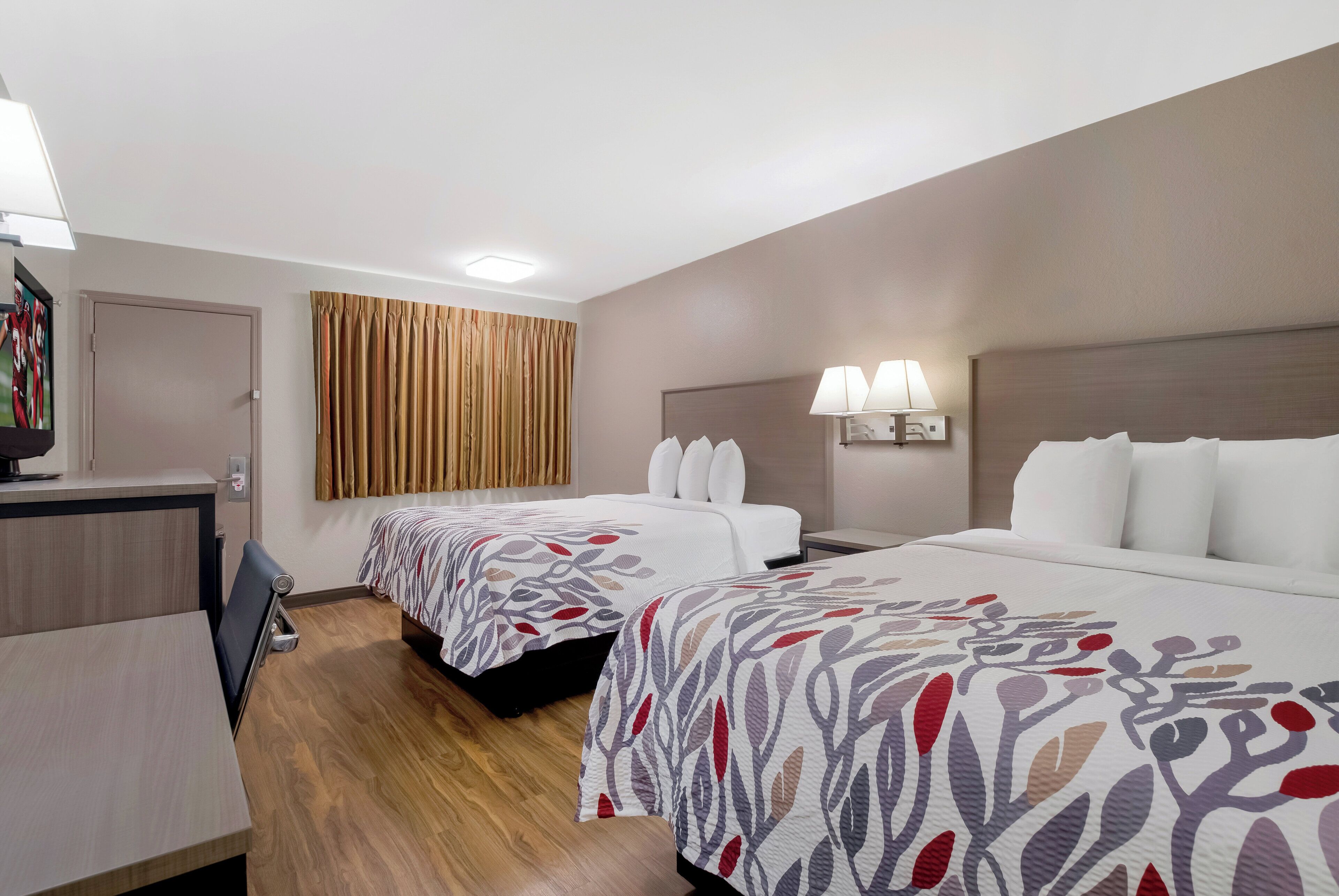 A hotel room with two beds, a desk, a chair, a TV, and a window with curtains.