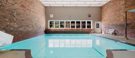 An indoor swimming pool with brick walls, a clear ceiling, and a window overlooking greenery.