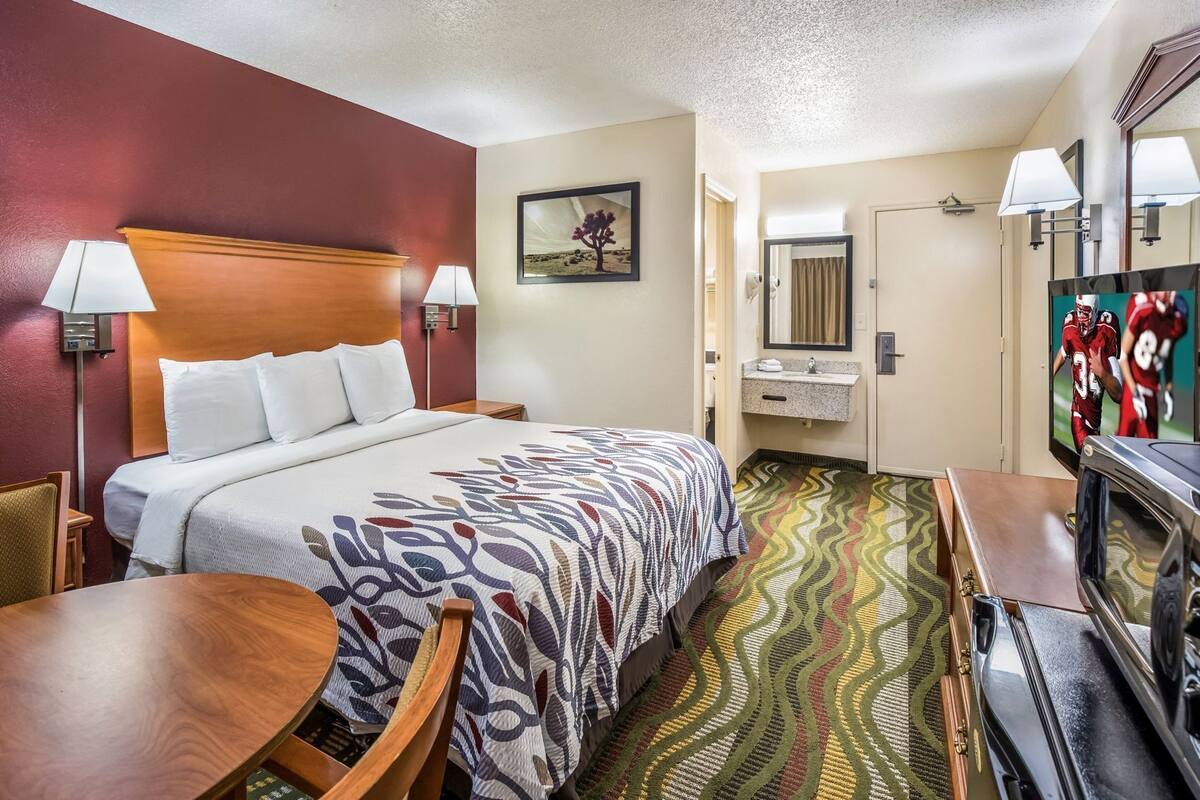 A hotel room with a bed, bedside tables, a desk, a television, a mirror, a sink, and a patterned carpet.