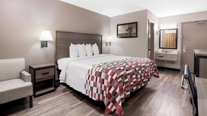 Una habitación de hotel con cama, una silla, una mesita de noche, lámpara, un cuadro, un baño con espejo y una puerta.