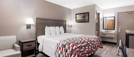 A hotel room with a bed, a chair, a nightstand, a bedside table, a lamp, a painting, a bathroom with a mirror, and a door.