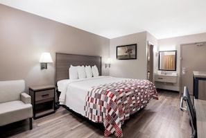 A hotel room with a bed, a chair, a nightstand, a bedside table, a lamp, a painting, a bathroom with a mirror, and a door. - Red Roof Inn Chattanooga - Lookout Mountain (Chattanooga)