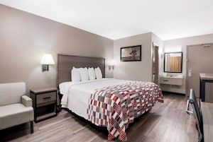 A hotel room with a bed, a chair, a nightstand, a bedside table, a lamp, a painting, a bathroom with a mirror, and a door.