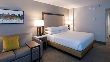 Una habitació d'hotel amb un llit, una tauleta de nit, una làmpada, un sofà amb un coixí groc i una obra d'art amb un paisatge urbà a la paret.
