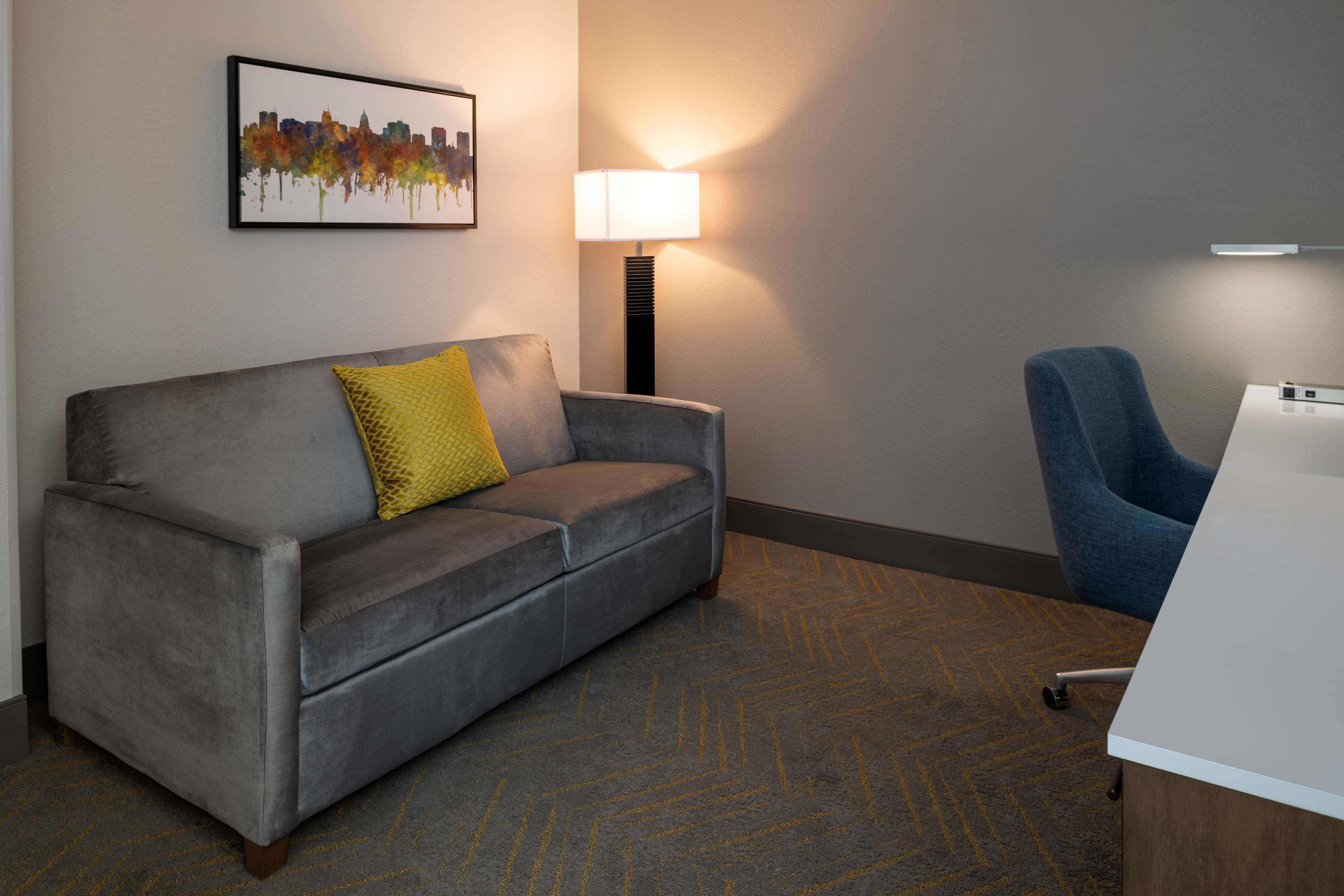 Une chambre d’hôtel moderne avec un canapé gris, un coussin jaune, une lampe sur pied, une chaise bleue et un bureau avec un ordinateur.