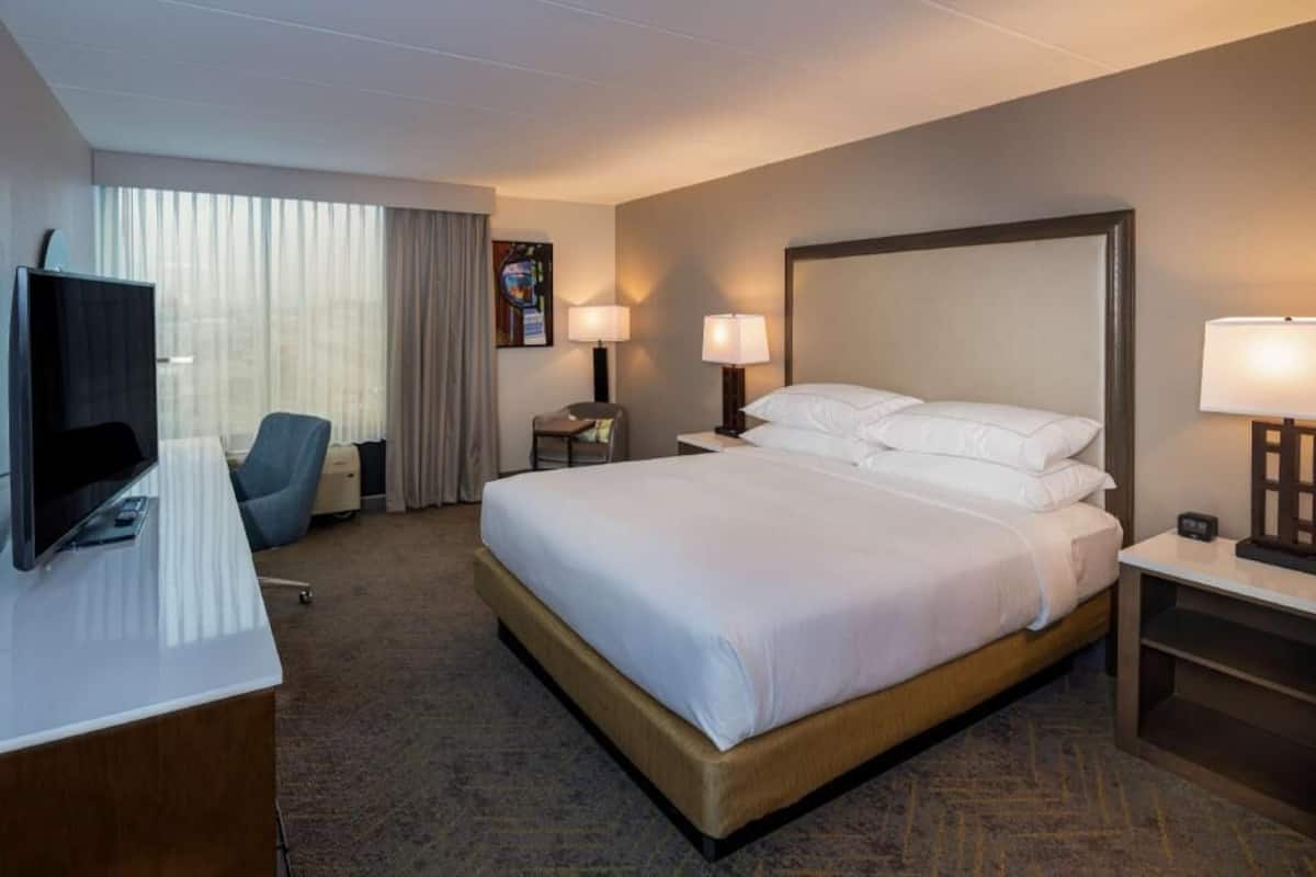 Una habitación de hotel con cama, escritorio, silla, televisor y ventana con cortinas.