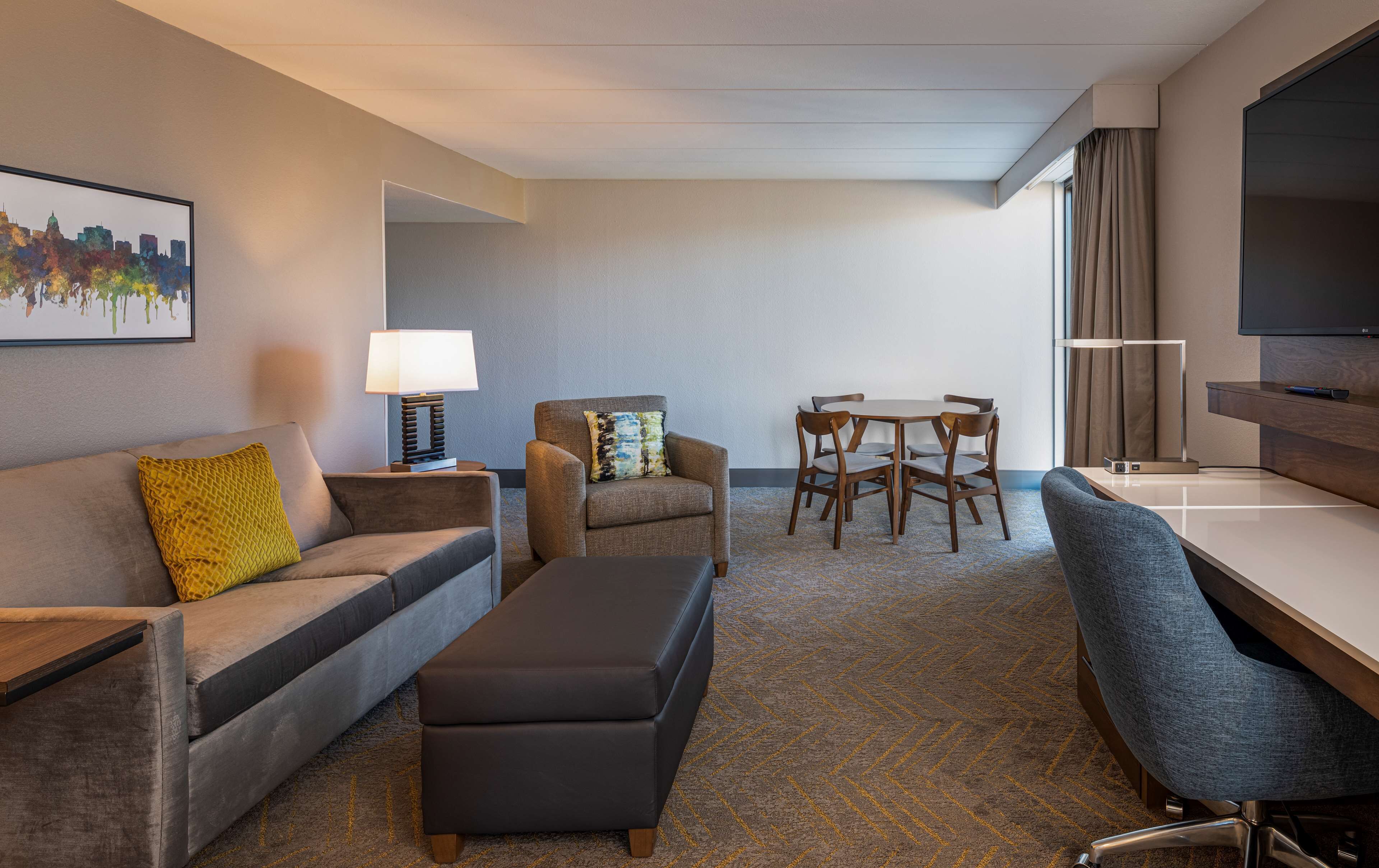 A hotel room with a sofa, ottoman, armchair, dining table, chairs, a TV, and a lamp.
